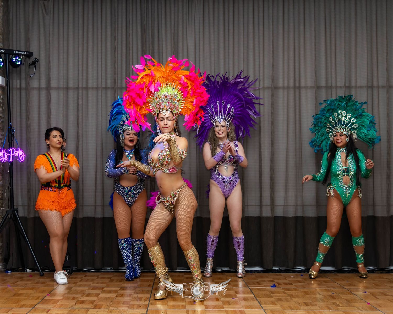 Lineup of performers in vibrant carnival costumes with feathered headdresses on stage.