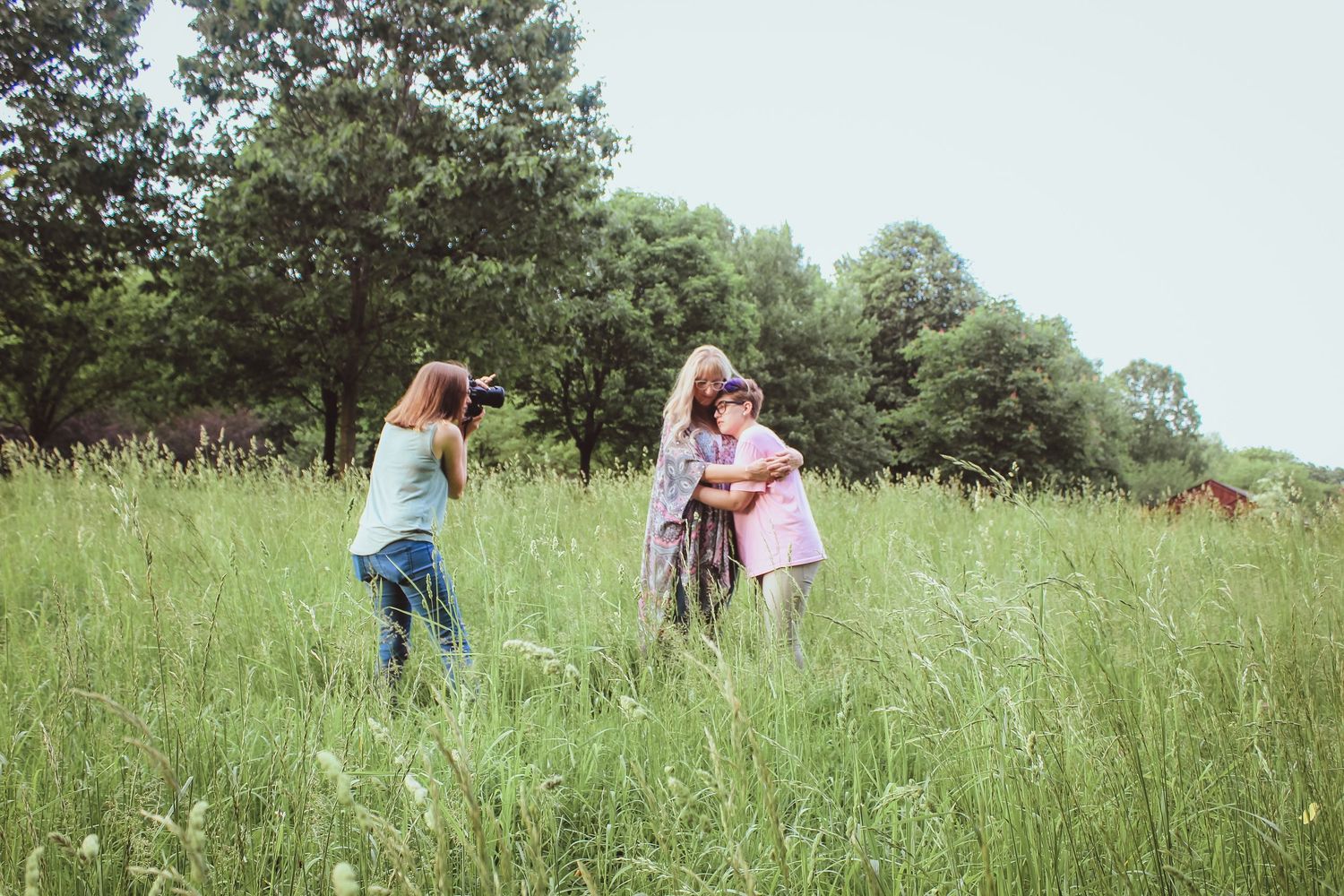 Two people pose for photos with one photographer in a lush summer meadow with tall grass and trees in the background.