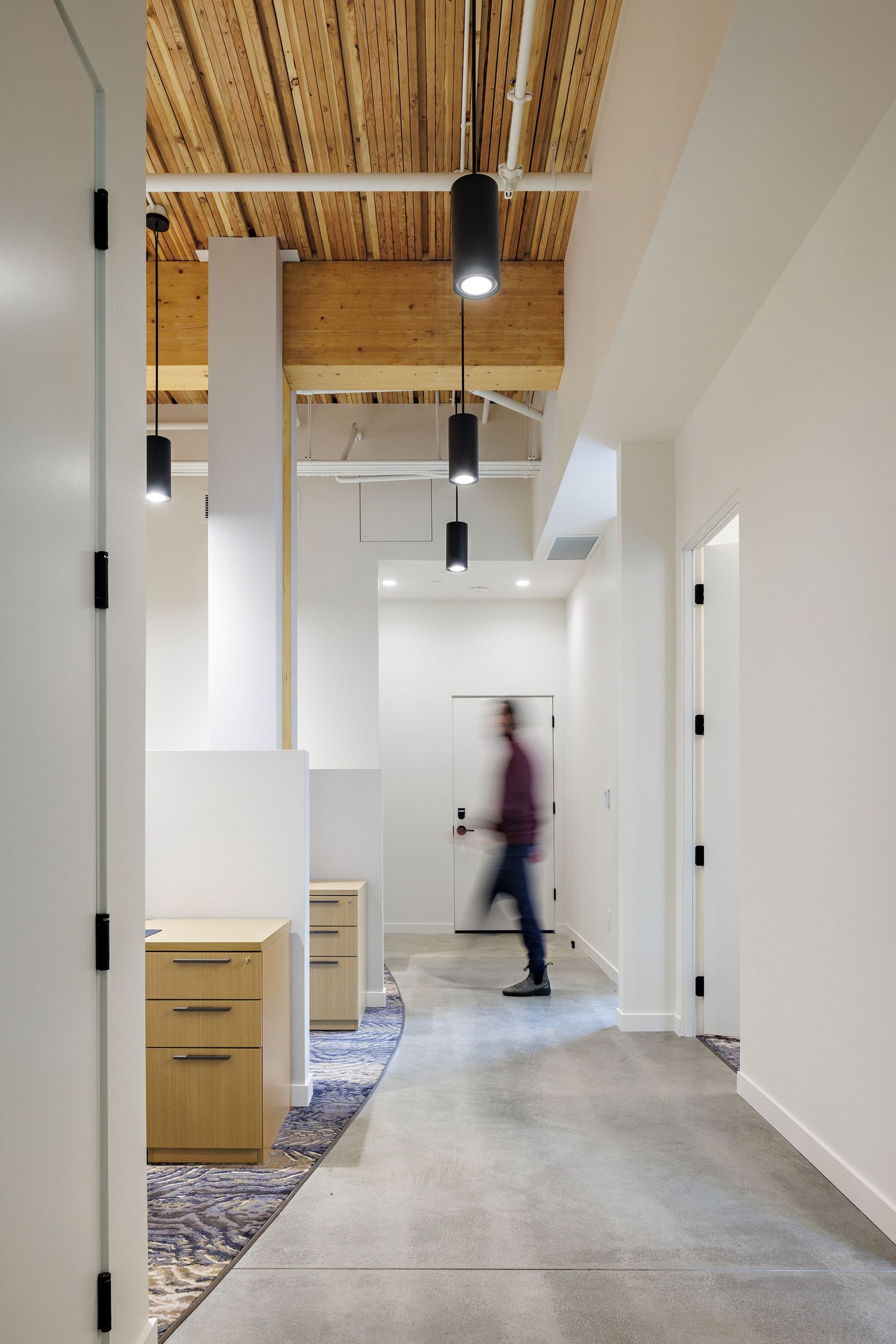 Modern office hallway with concrete floors, wood ceiling, pendant lights and blurred moving figure walking past white walls.