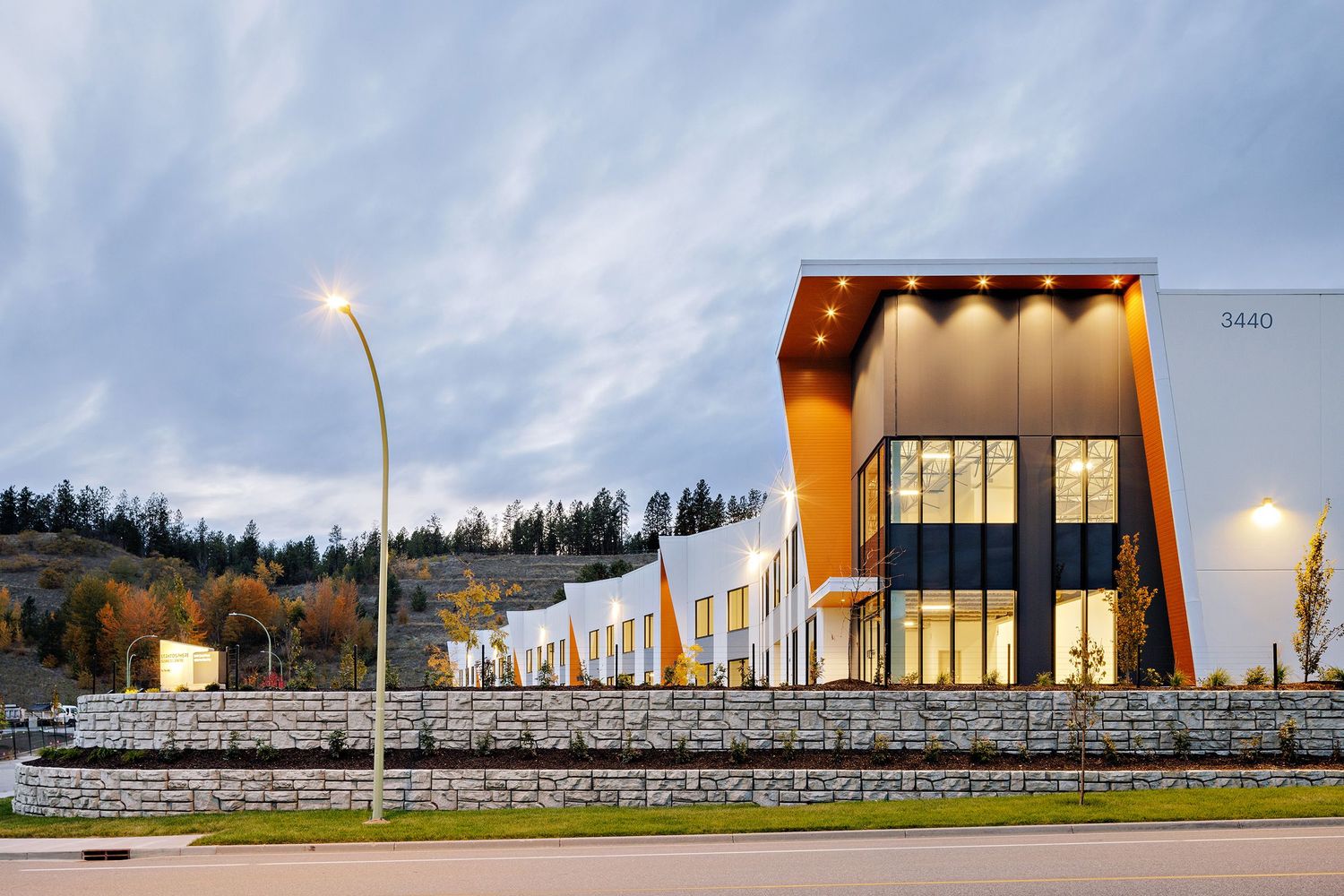 Modern curved commercial building with glass windows and stone facade illuminated at dusk under cloudy skies.