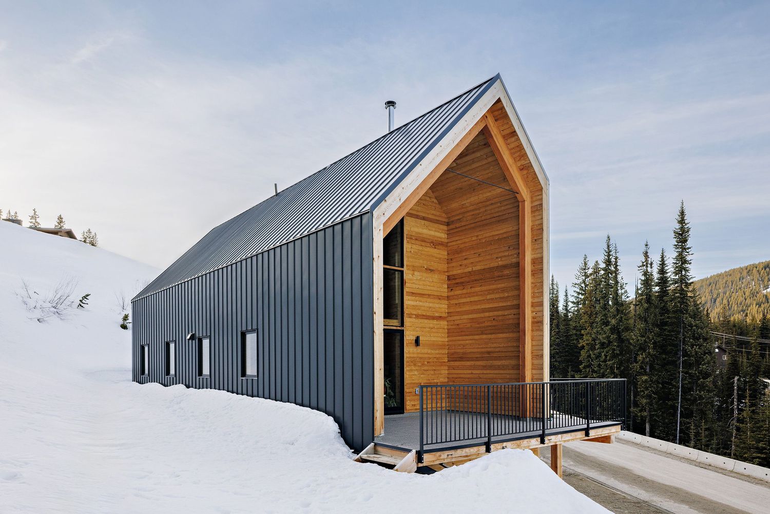 Modern mountain cabin with grey metal siding and wood accents sits nestled in snowy landscape among evergreen trees.