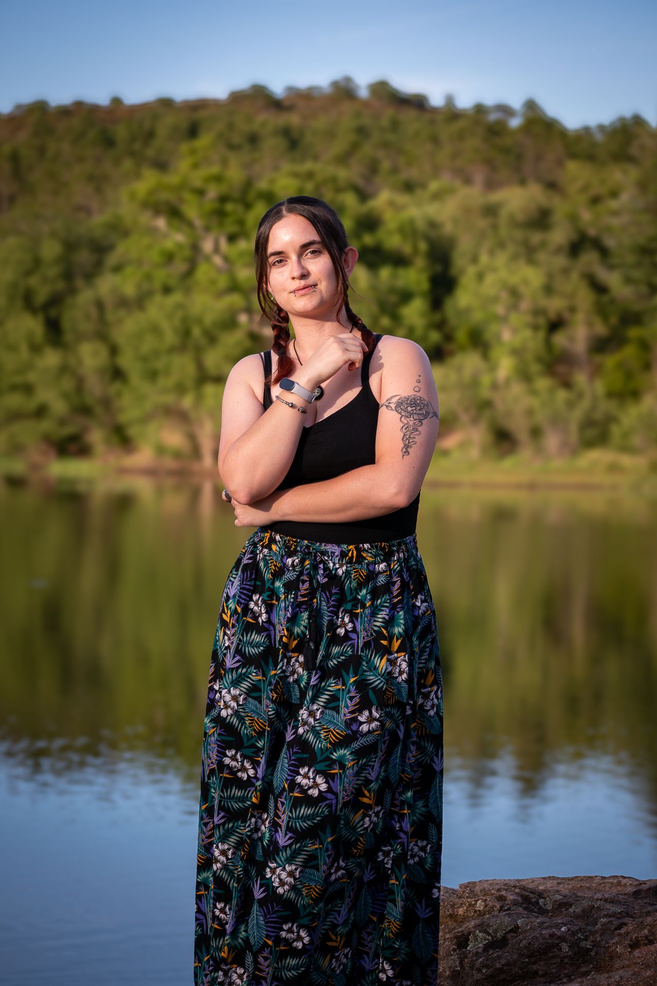 Person in a black top and floral print maxi skirt poses confidently by a serene lake with green hills in background.