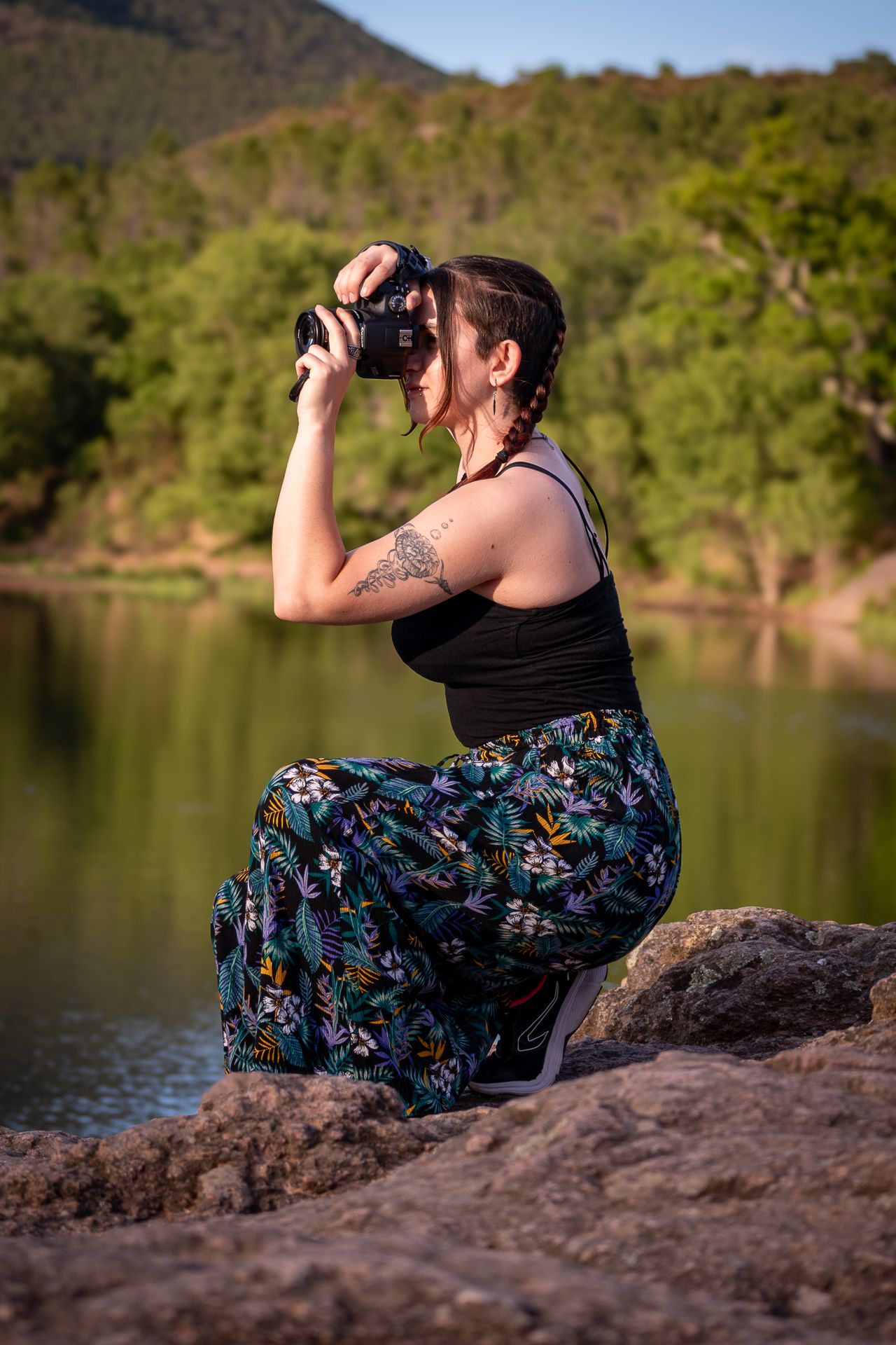 Photographer in floral skirt and black tank top takes photos while sitting on rocks by a serene lake at sunset.