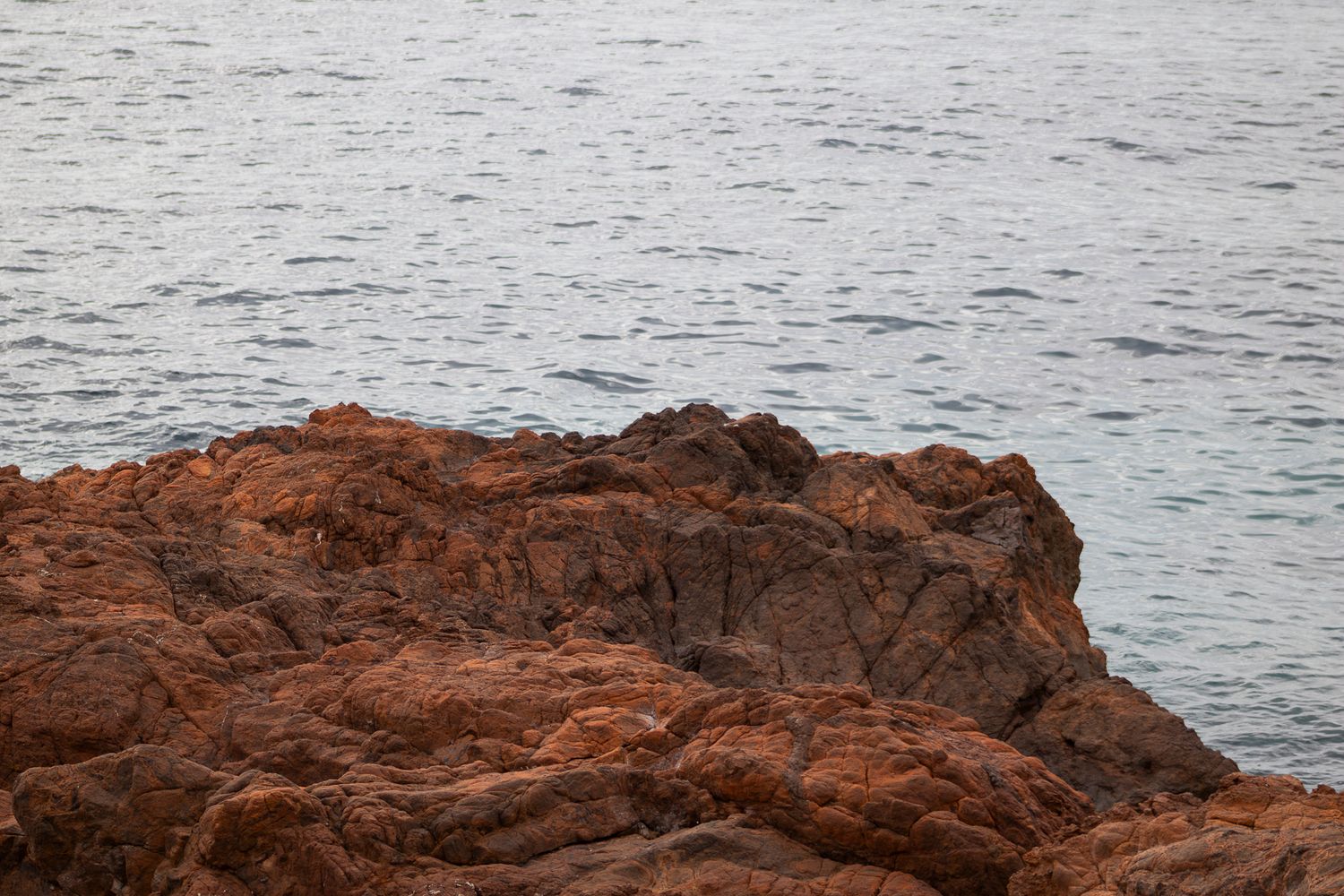 Red rocky coastal formation extends into rippling ocean waters against a gray seascape.