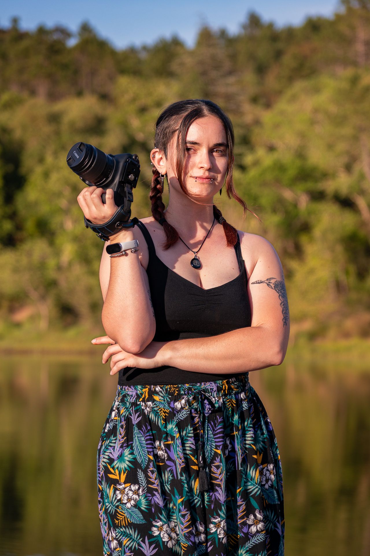 Photographer in black top and floral skirt stands near lake at golden hour holding professional camera.