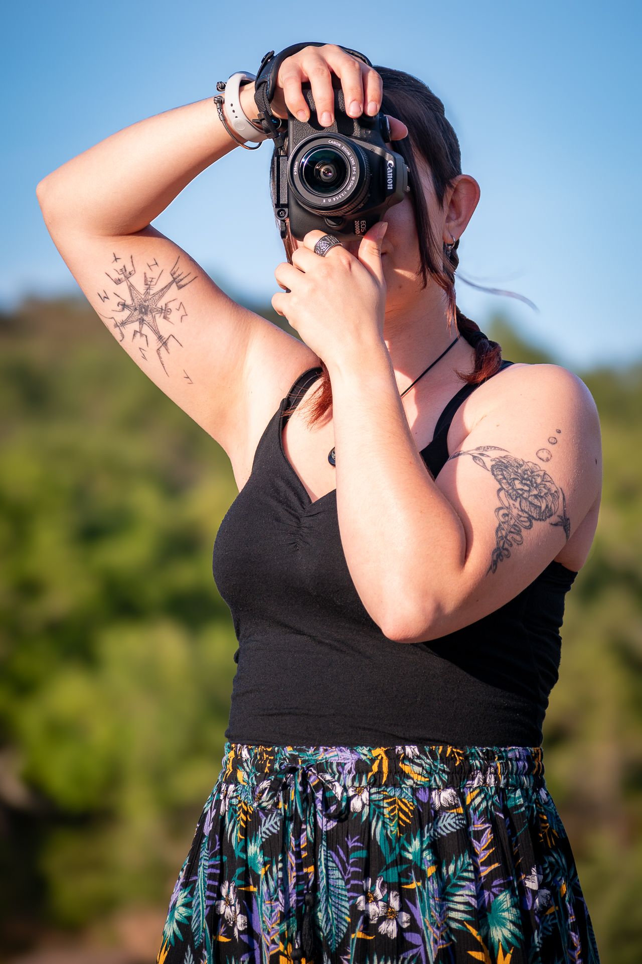 Photographer wearing black tank top and floral skirt takes photo with DSLR camera against blurred nature background.