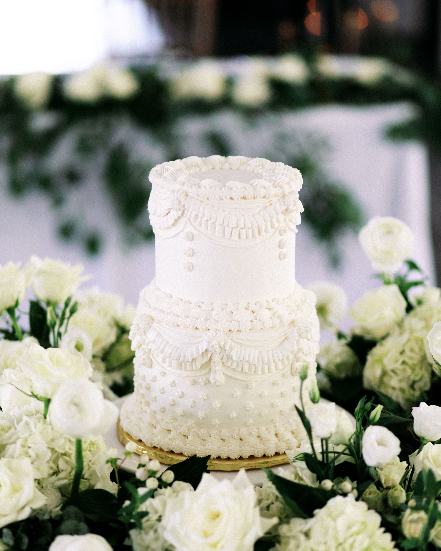 Elegant white wedding cake decorated with lace details surrounded by white roses.
