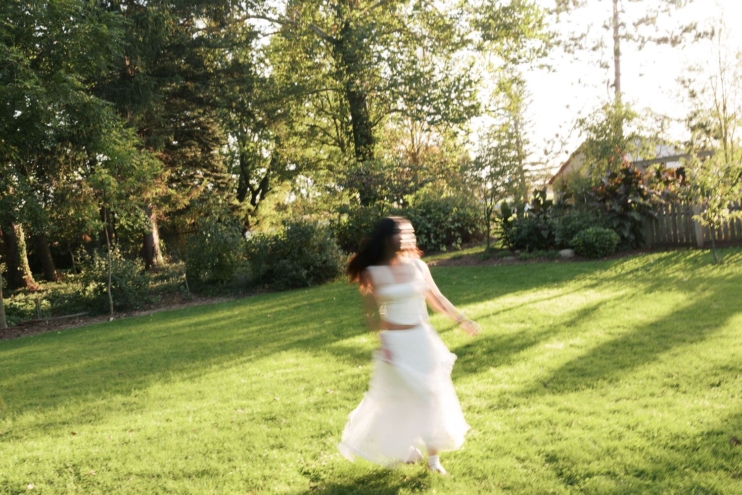 A person in a flowing white dress twirls joyfully in a sunlit garden setting.