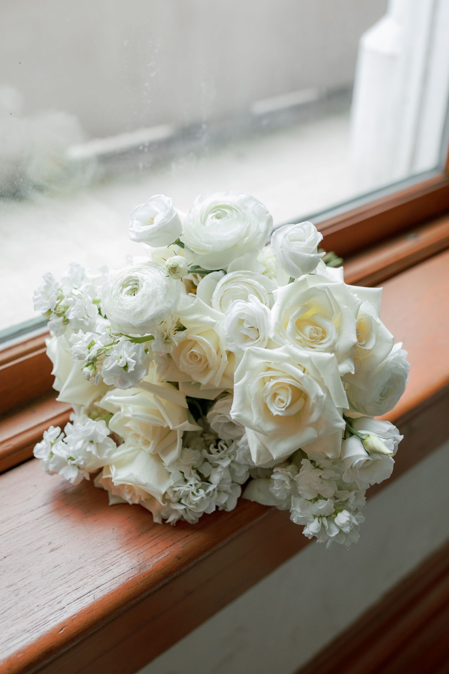 Elegant white rose and ranunculus bridal bouquet resting on wooden windowsill.