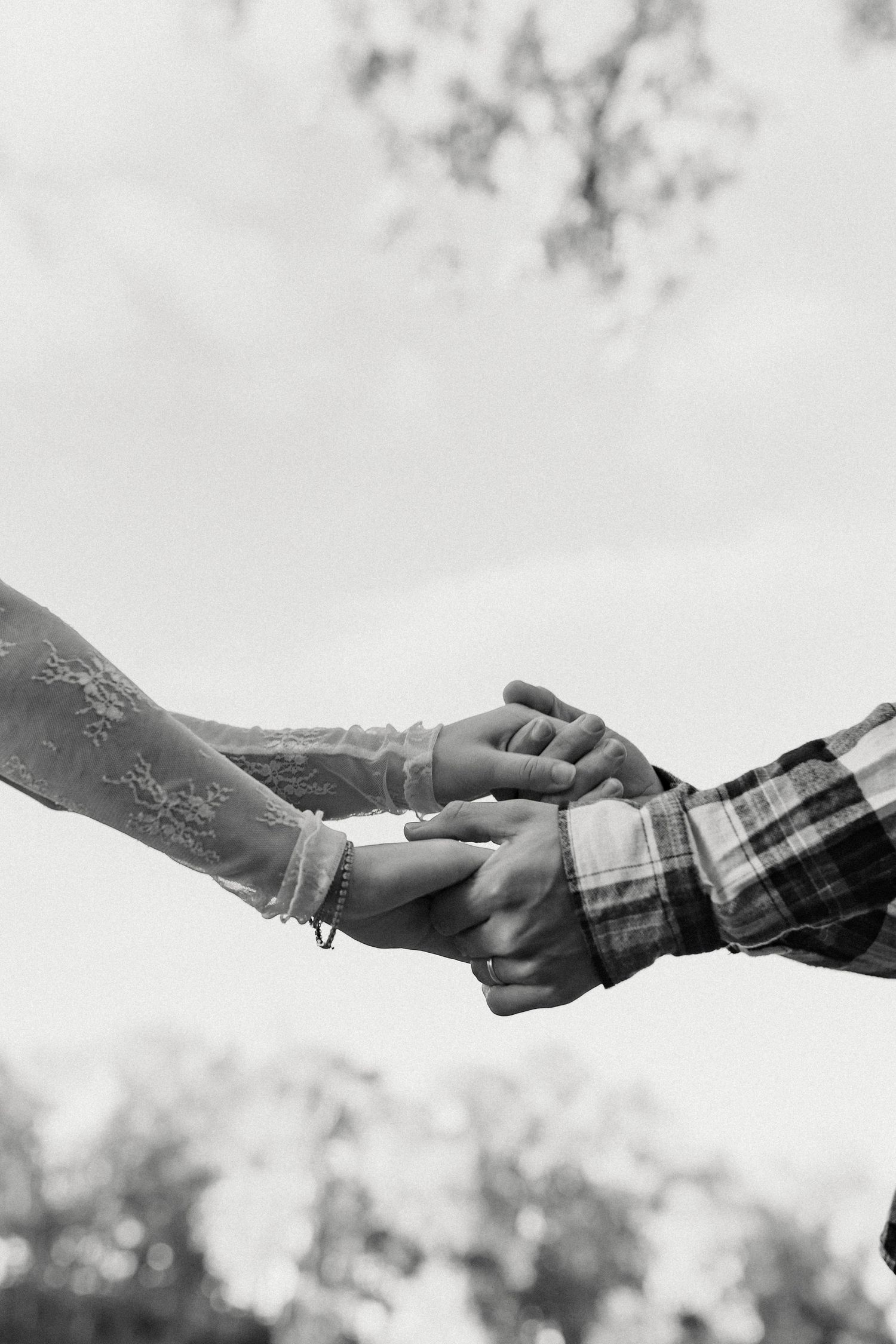 Black and white close-up of two hands reaching out to hold each other against a blurred sky.