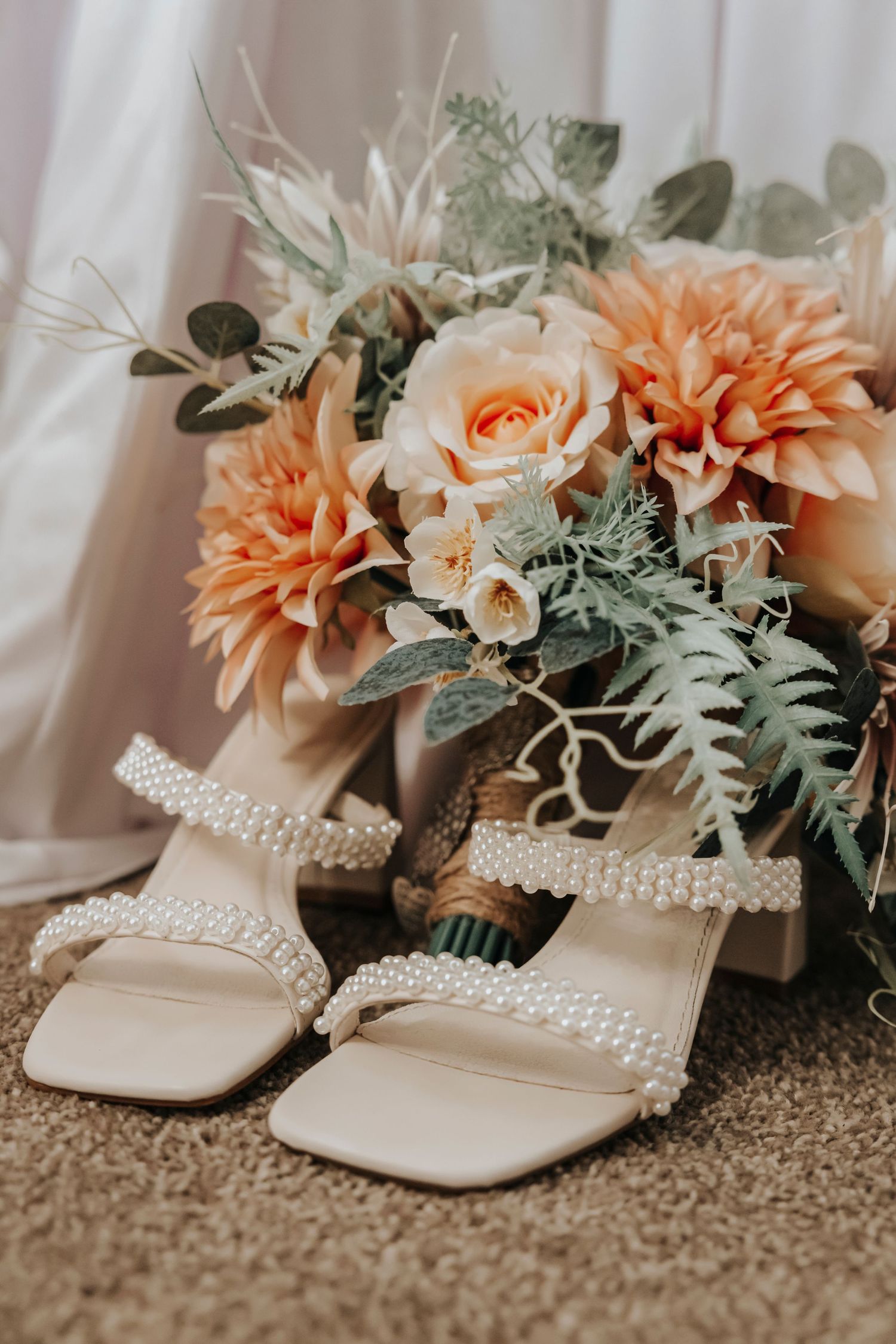 Pearl embellished bridal sandals next to a peachy floral bouquet with roses, dahlias and eucalyptus on neutral fabric.