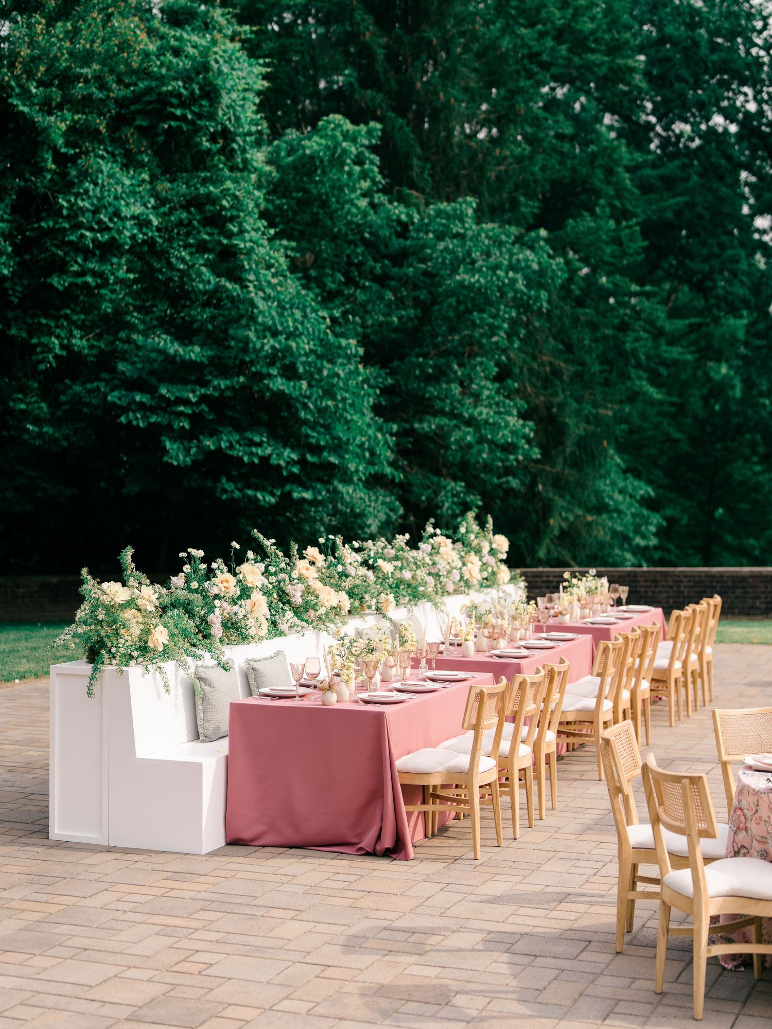 Elegant outdoor wedding reception setup with dusty rose tablecloths, wooden chairs and lush white floral arrangements.