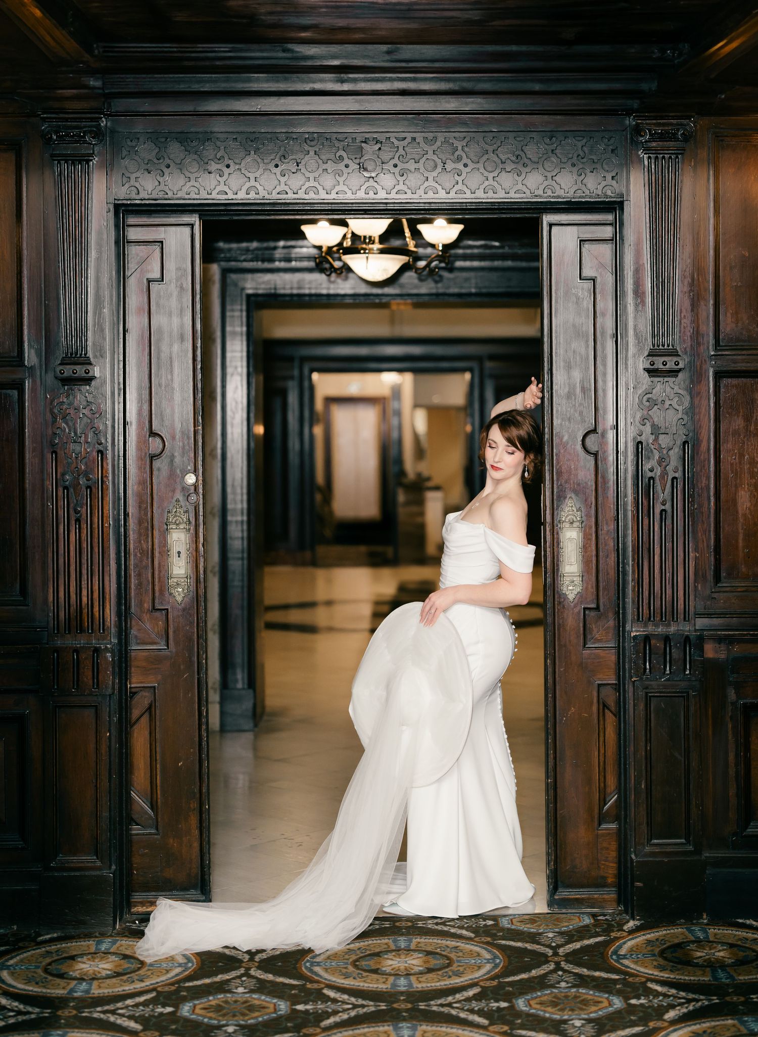 A bride in a flowing white wedding dress stands elegantly in an ornate dark wood doorway with vintage architectural details.