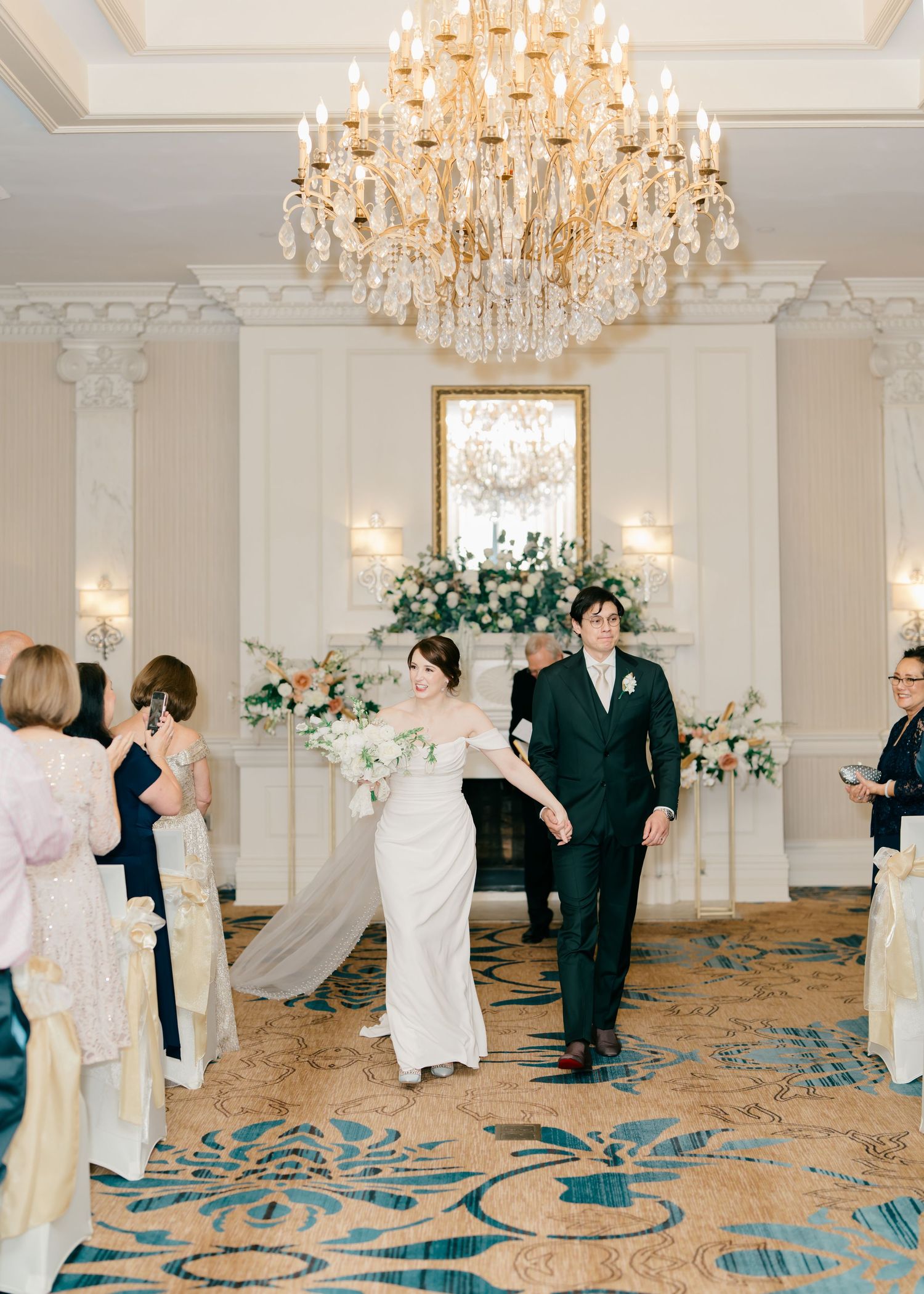 Elegant wedding ceremony in a grand ballroom with crystal chandelier and floral arrangements against white walls.