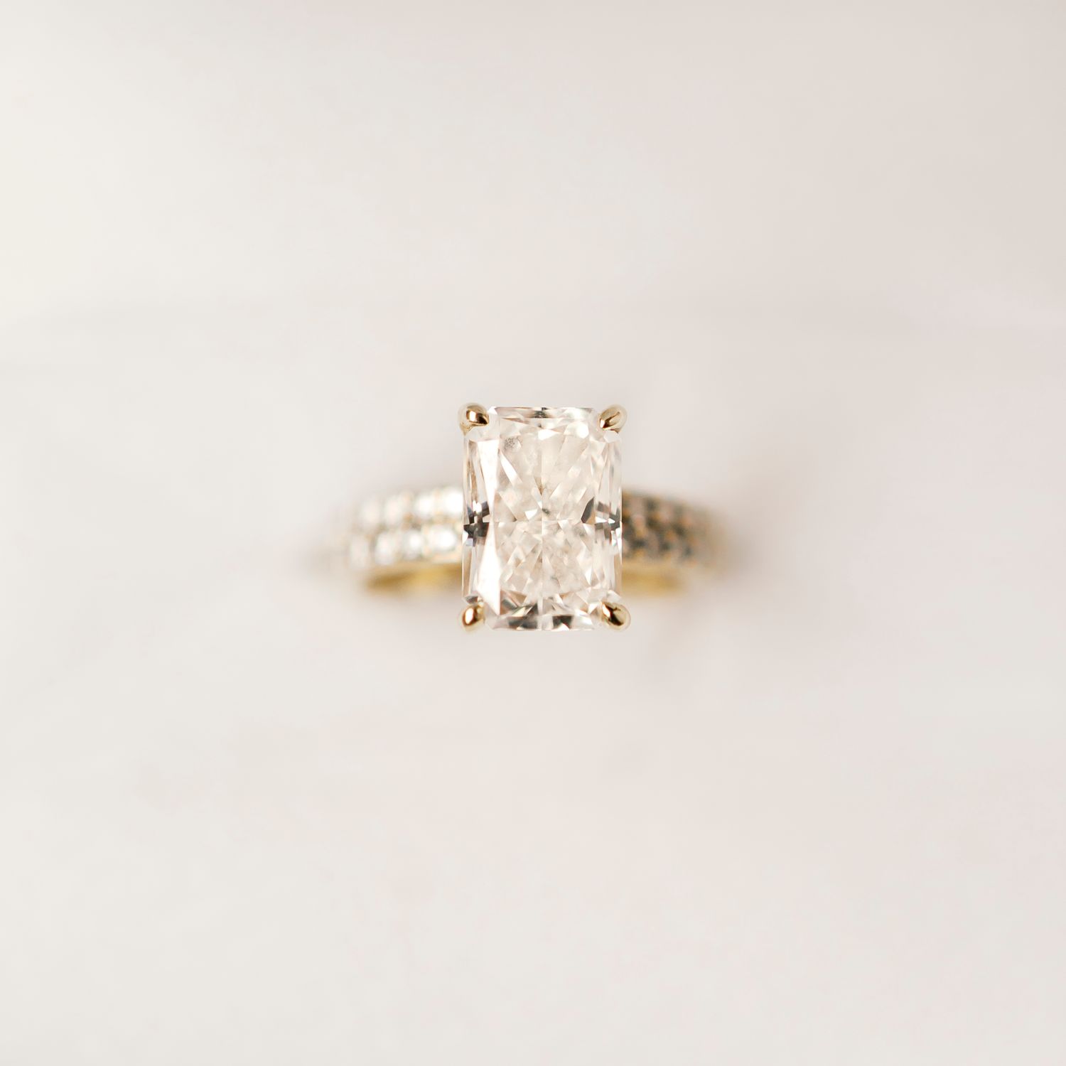 An elegant engagement ring featuring a radiant cut diamond on a gold band, photographed against a soft beige background.