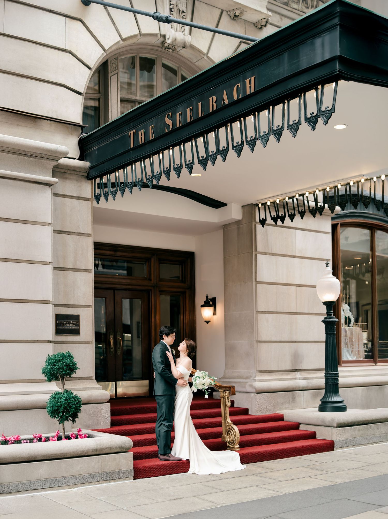 Elegant couple stands on red-carpeted steps outside The Selfbach luxury building entrance with ornate green awning.