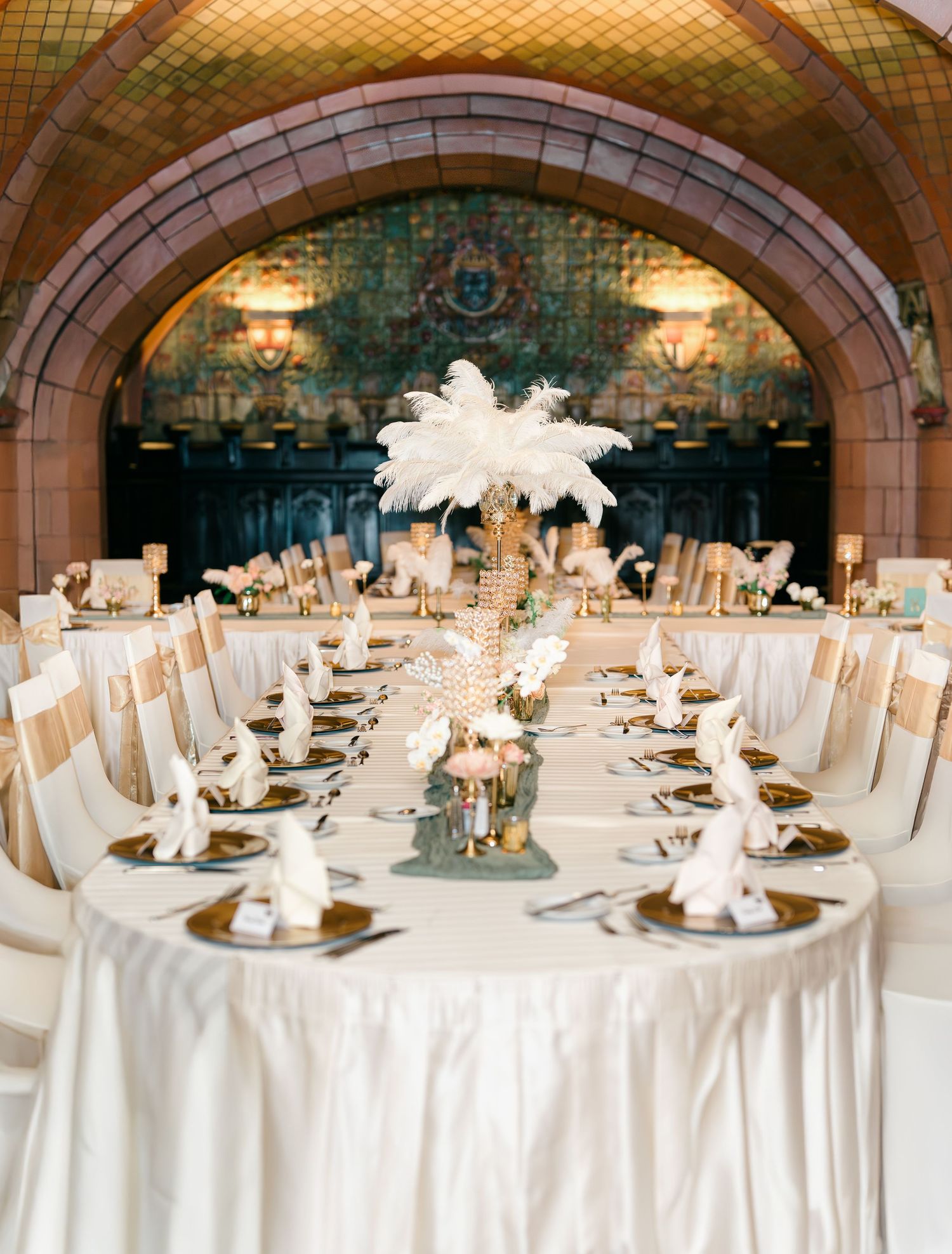 Elegant wedding reception tables with white linens and ornate centerpieces featuring feathers in an arched ballroom setting.