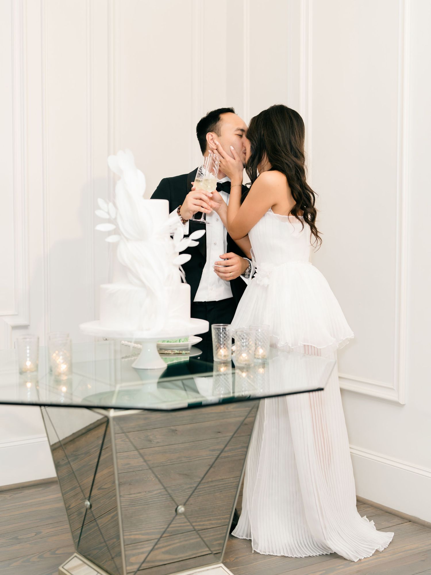 Newlywed couple shares intimate moment while toasting with champagne glasses beside modern glass table with candles.