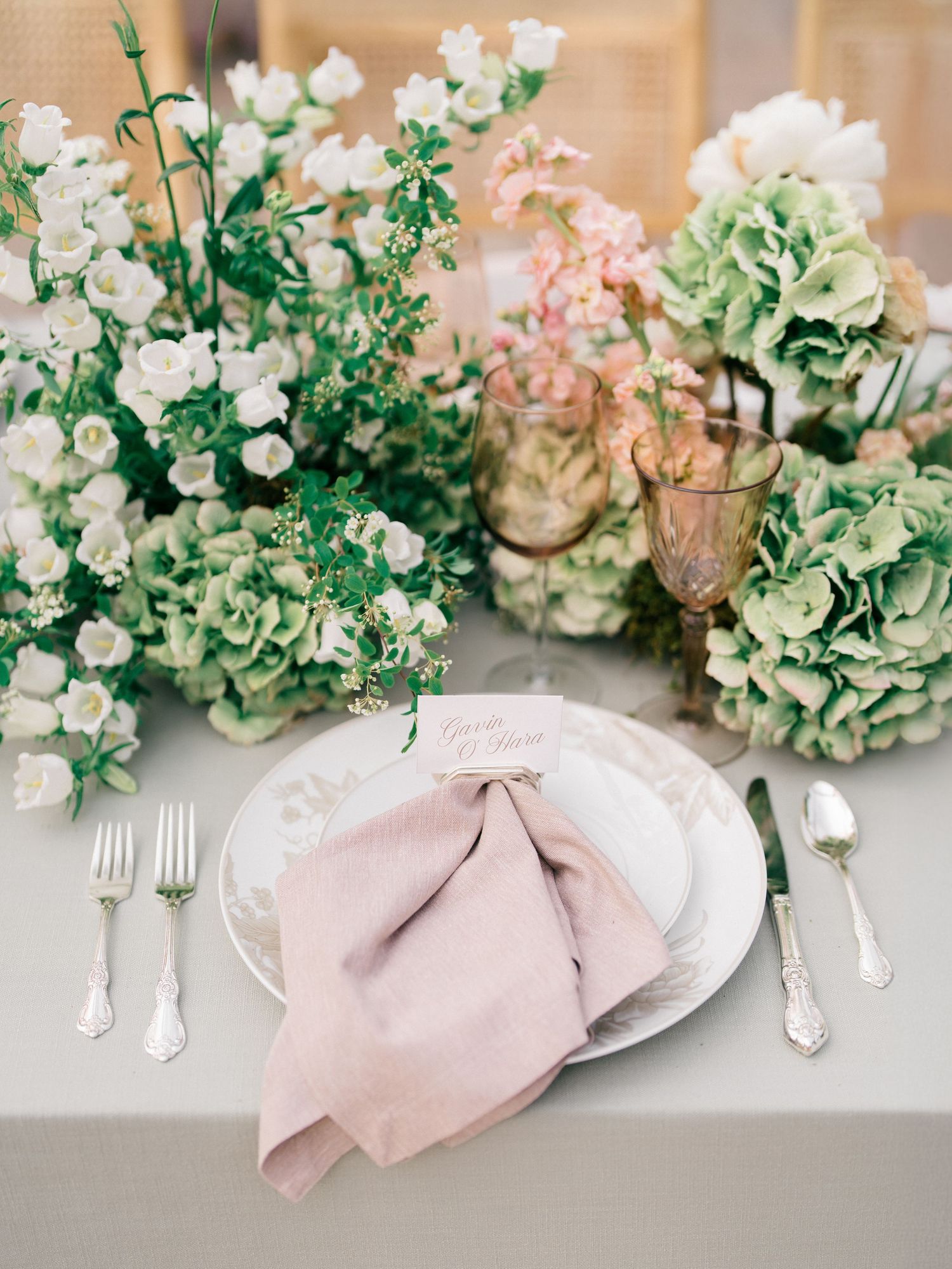 Elegant table setting with blush pink napkin and white flowers against soft gray linen tablecloth and vintage silver cutlery.