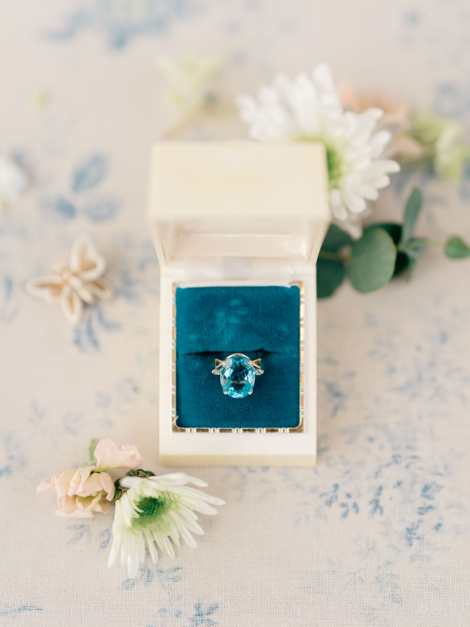 Aqua blue engagement ring displayed in white velvet ring box with white flowers and eucalyptus on light blue background.