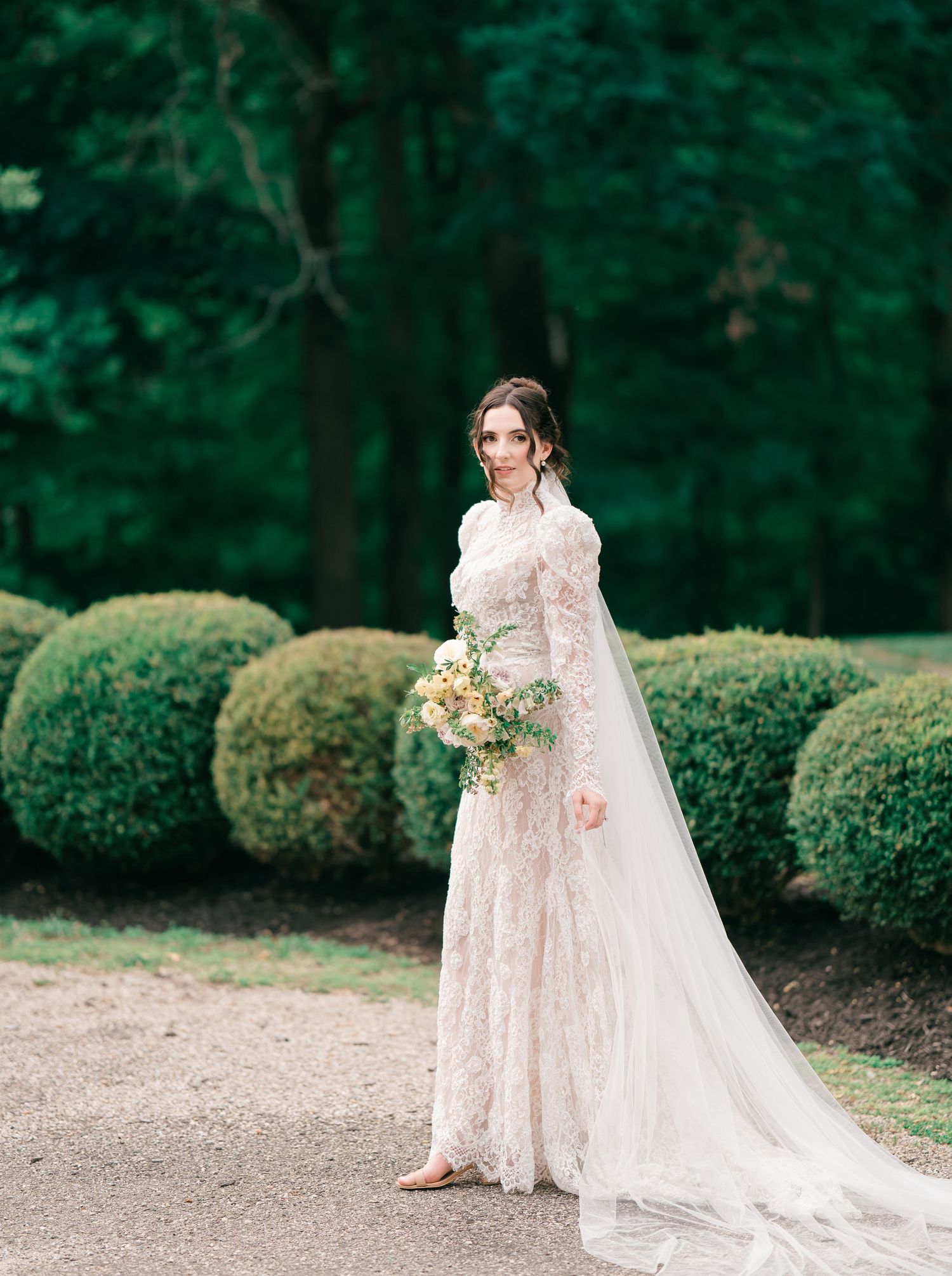 A bride in a long-sleeved lace wedding gown and veil stands elegantly along a path lined with manicured boxwood spheres.