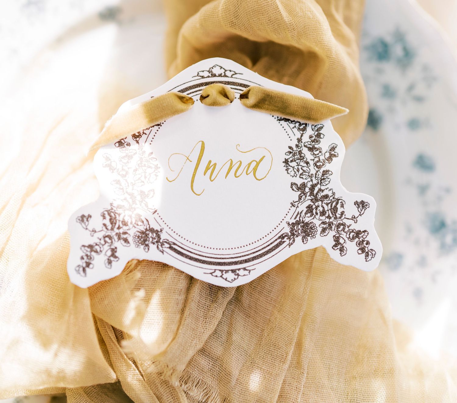 Elegant gold and white wedding place card with 'Anna' name and delicate floral decorative details.