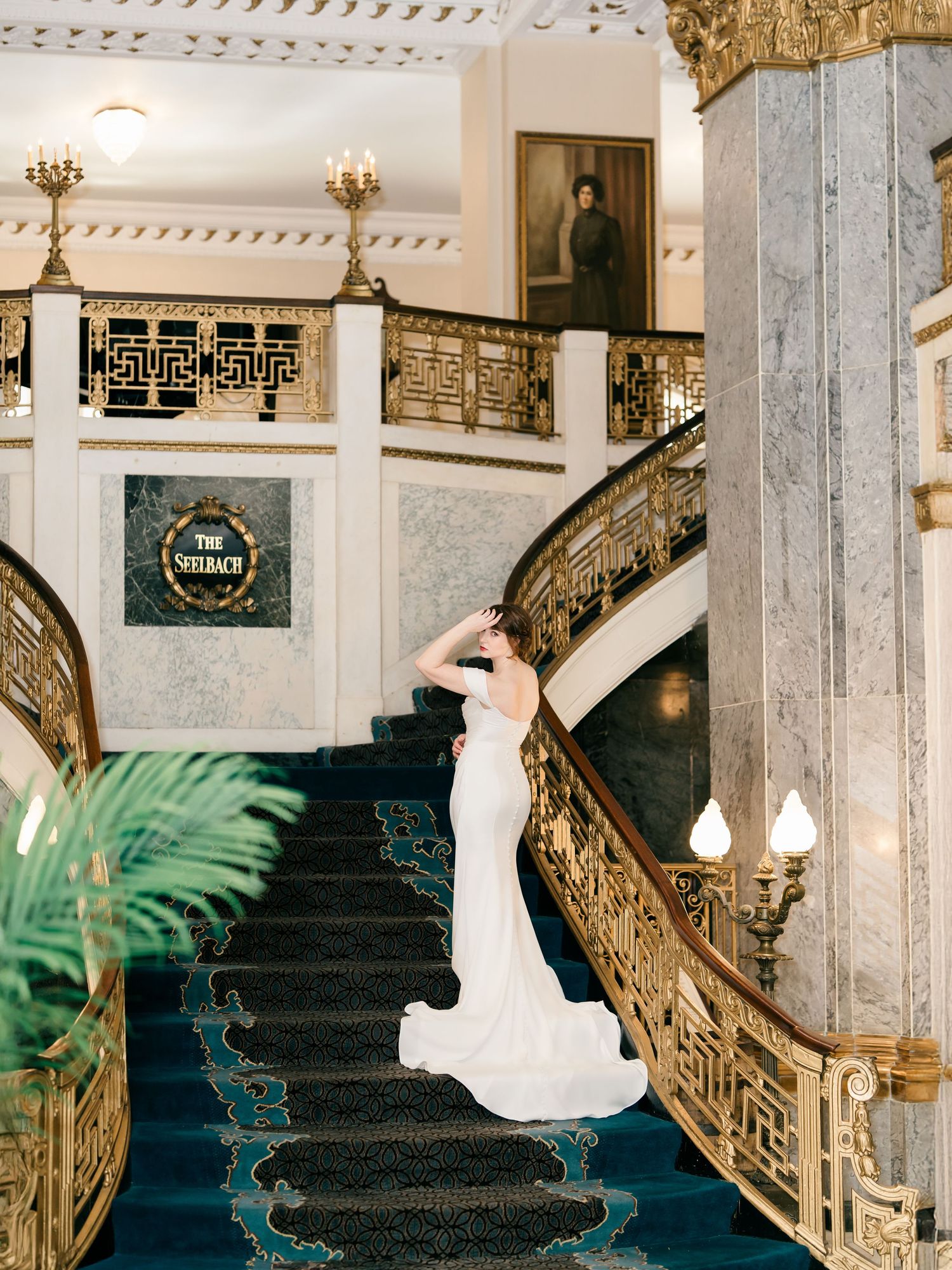 Elegant bride in white gown poses on grand staircase with ornate gold railings in historic hotel interior.