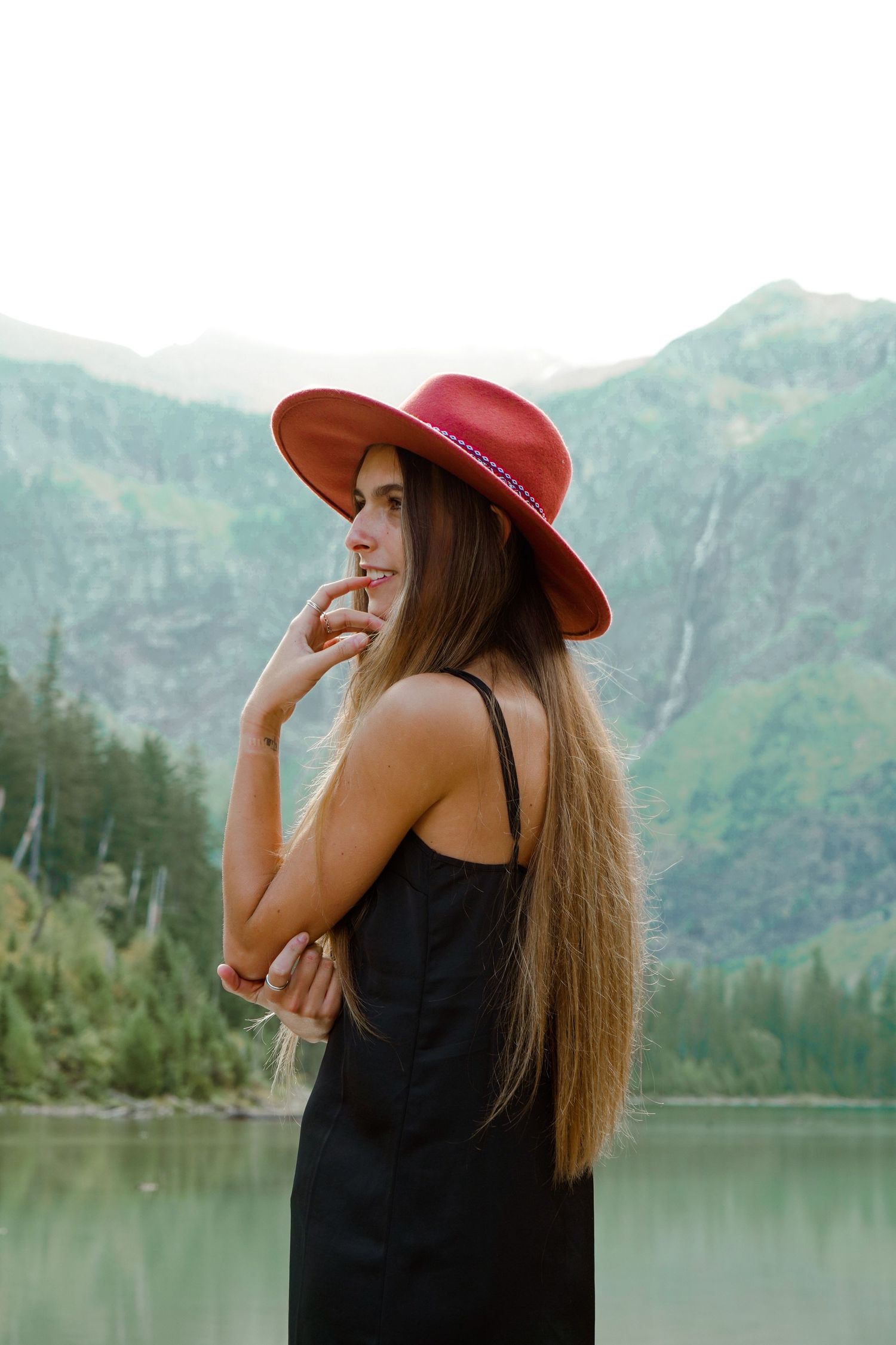 Person in black dress and red hat stands thoughtfully by mountain lake with scenic waterfall view in background.