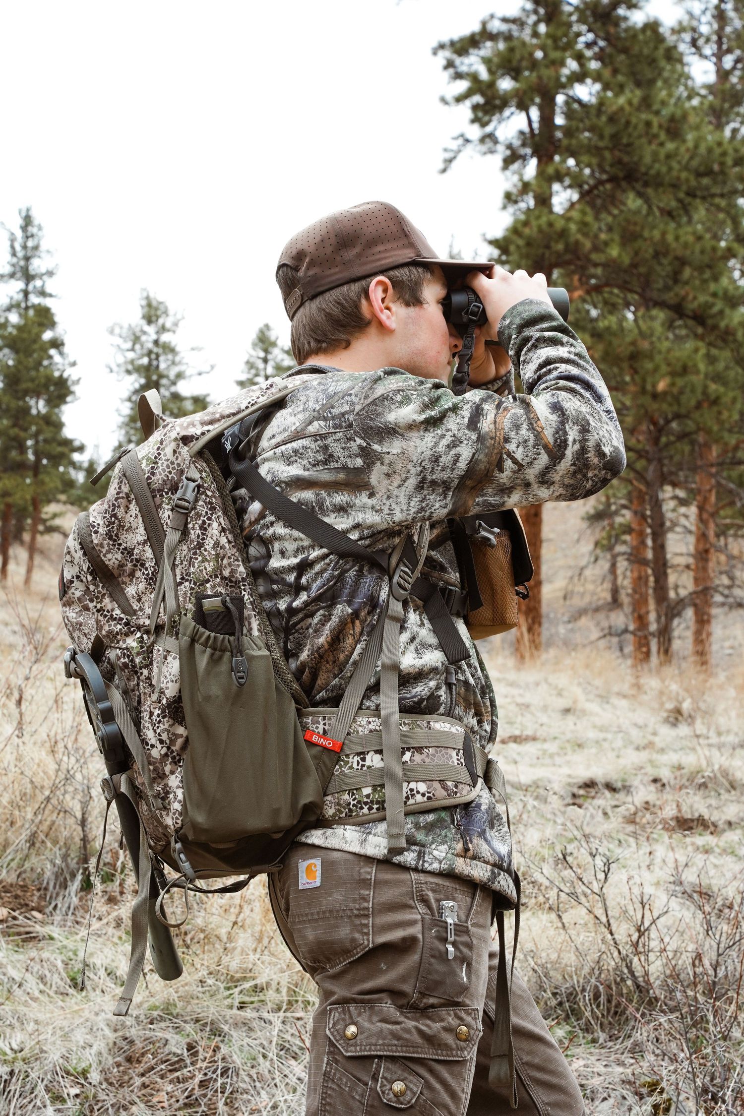 Hunter in camouflage gear using binoculars to scout terrain in a wooded area with backpack and hunting equipment.