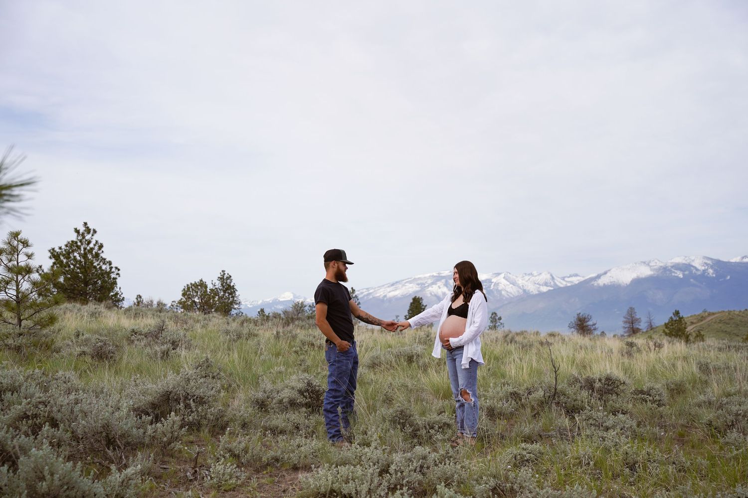 Two people stand together on a grassy hill with snow-capped mountains in the distance during an outdoor engagement.