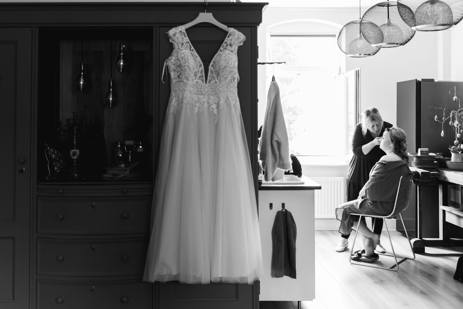 Getting Ready: Brautkleid hängt im Vordergrund, Braut wird im Hintergrund gestylt