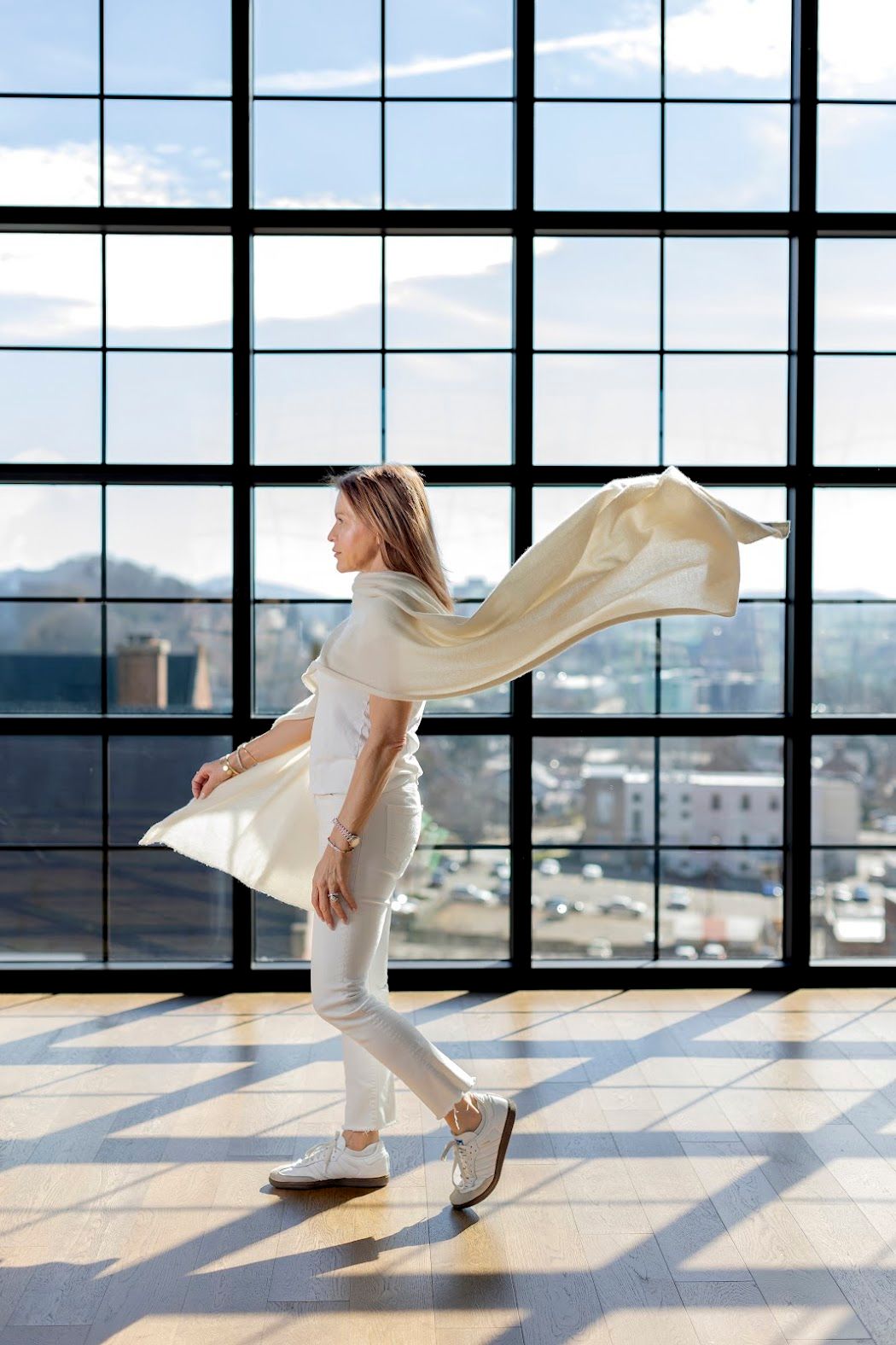 Tara in flowing white outfit walks in a sunlit studio with city of Asheville view through large windows.