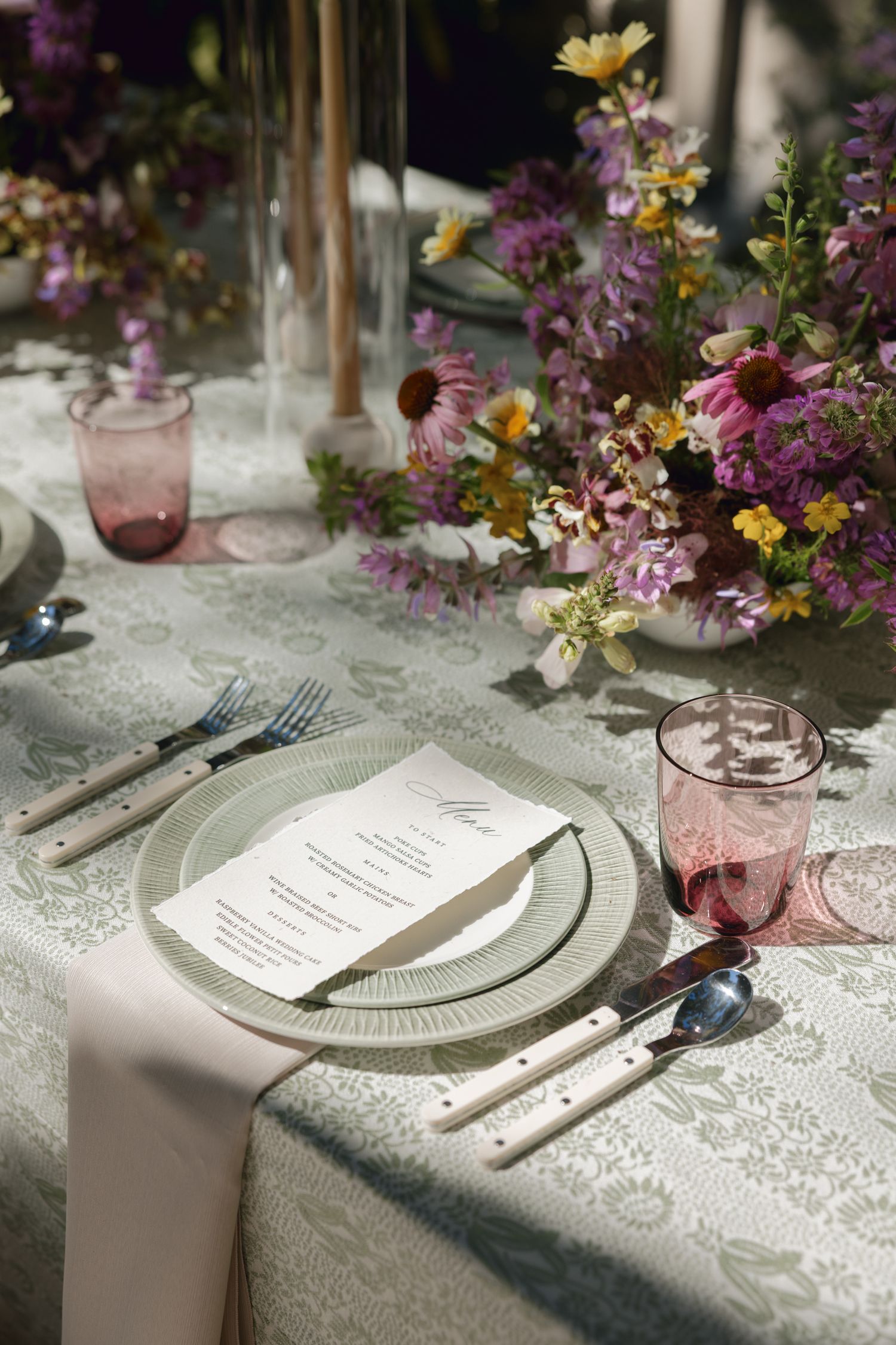 Elegant table setting with pink glassware, white plates, and purple floral arrangements on lace tablecloth.