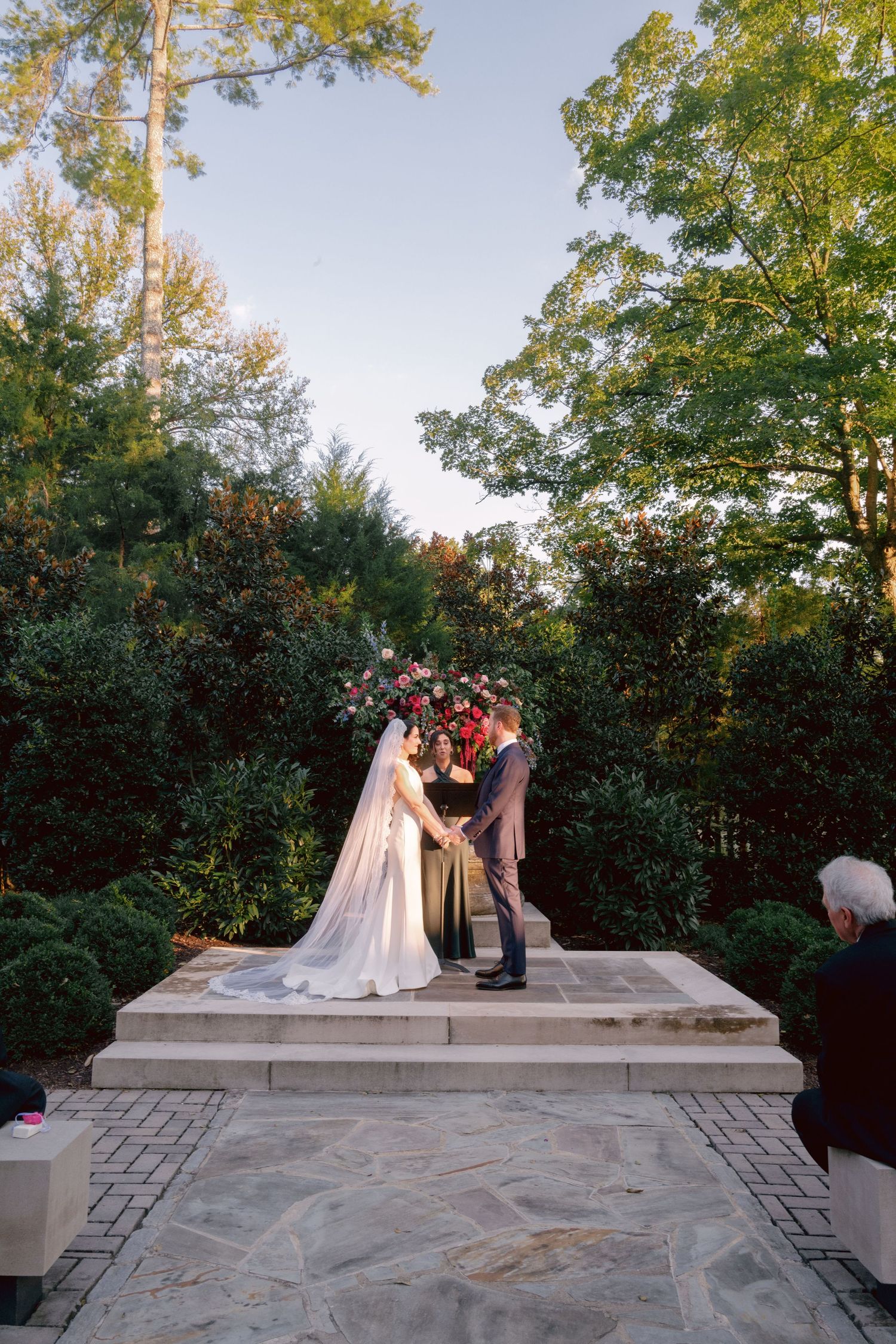 Wedding ceremony takes place on a stone terrace at Belle Meade Historic Site & Winery
surrounded by lush trees during golden hour.
