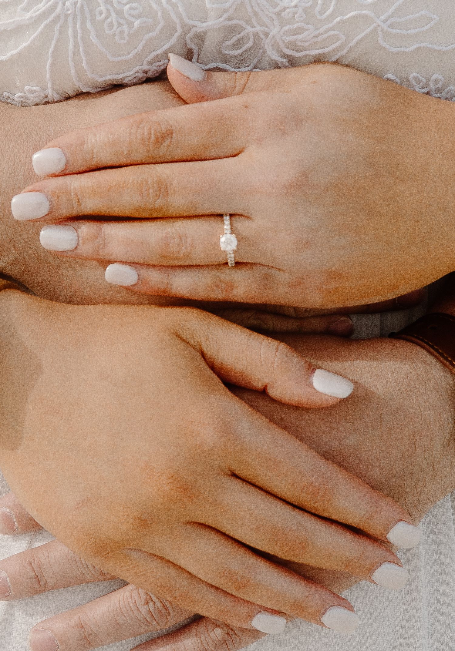Closeup of manicured hands with white nail polish displaying an elegant engagement ring against lace fabric.