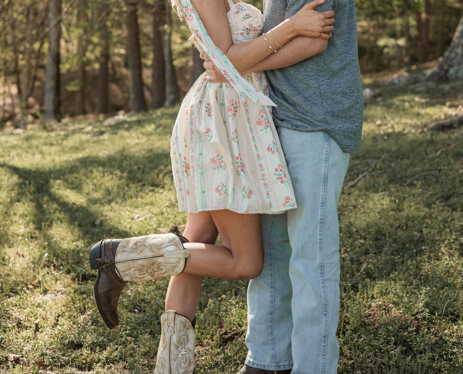 Romantic close-up shot showing legs in a floral dress and jeans embracing outdoors in a wooded setting.