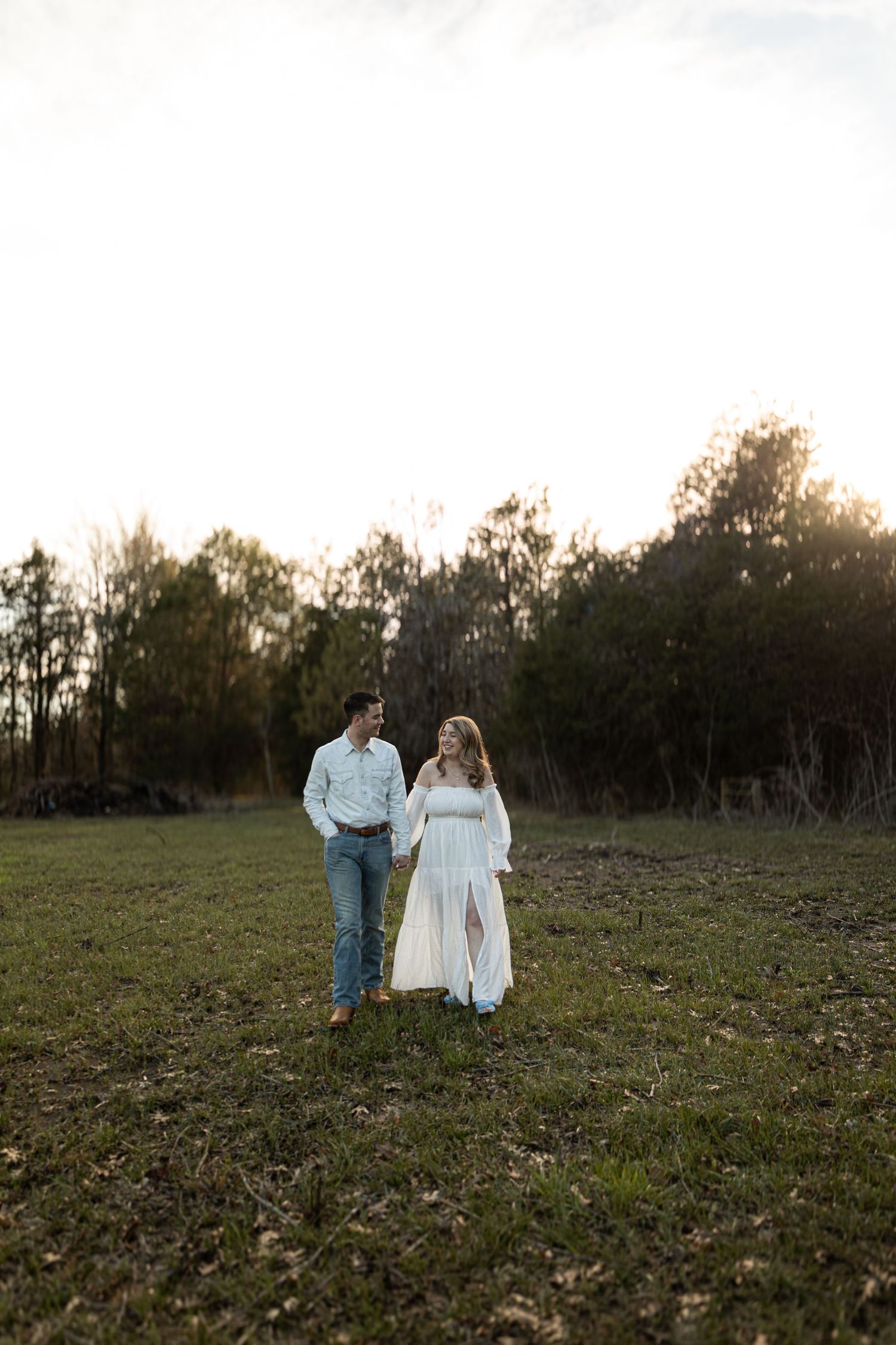 Couple walking together in a rustic field at sunset wearing white dress and denim.