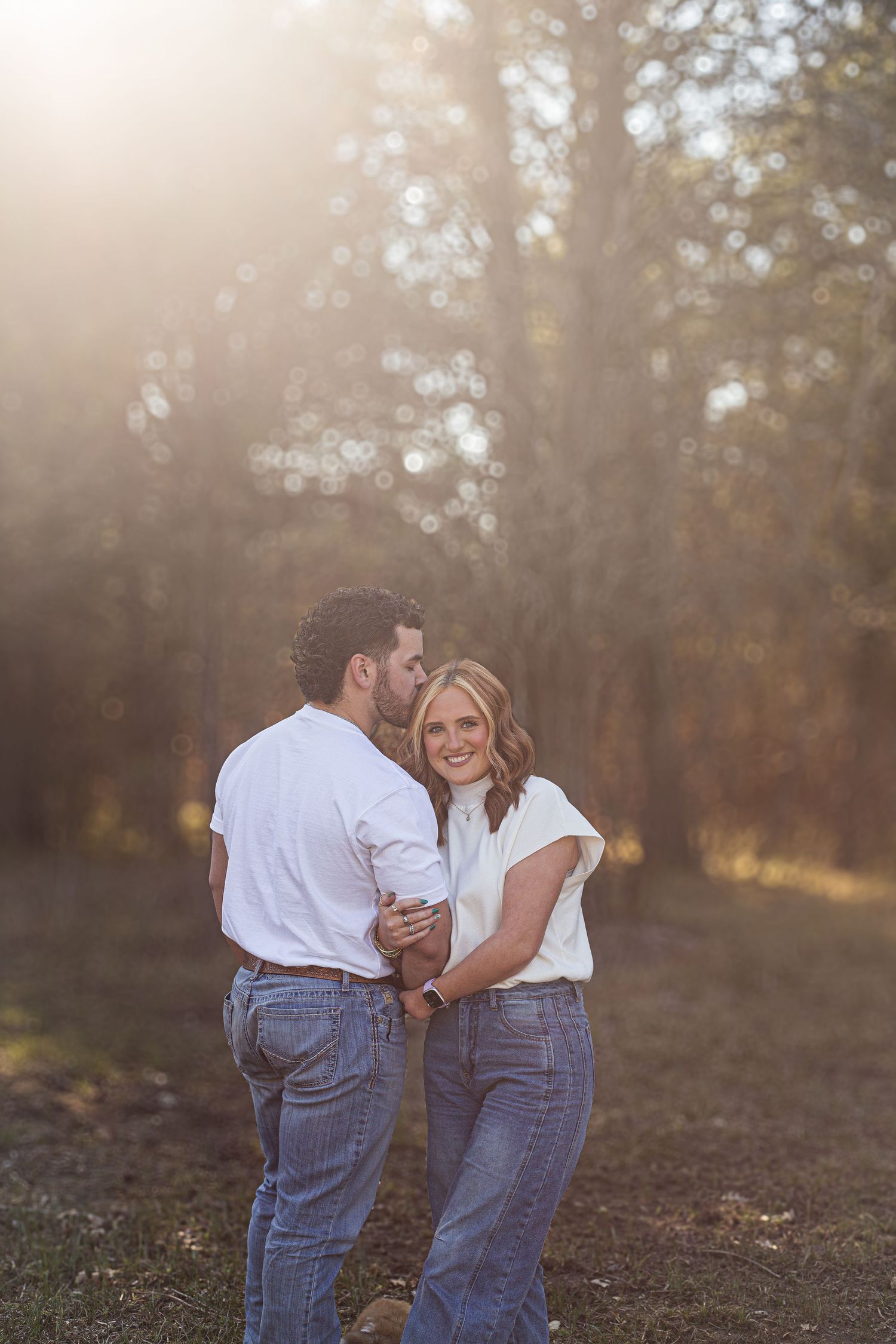 Couple in matching white shirts and jeans sharing a joyful embrace in a sunlit forest setting at golden hour.