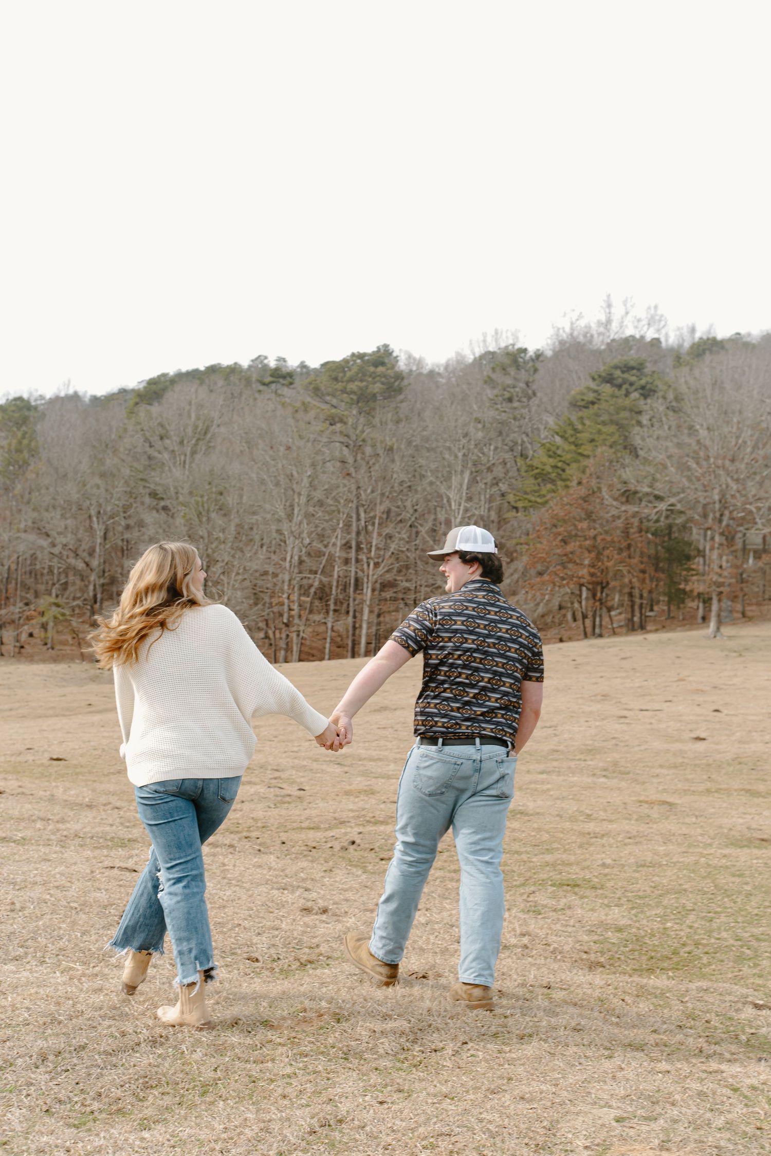 Couple holding hands while walking through an open field with trees in the background.