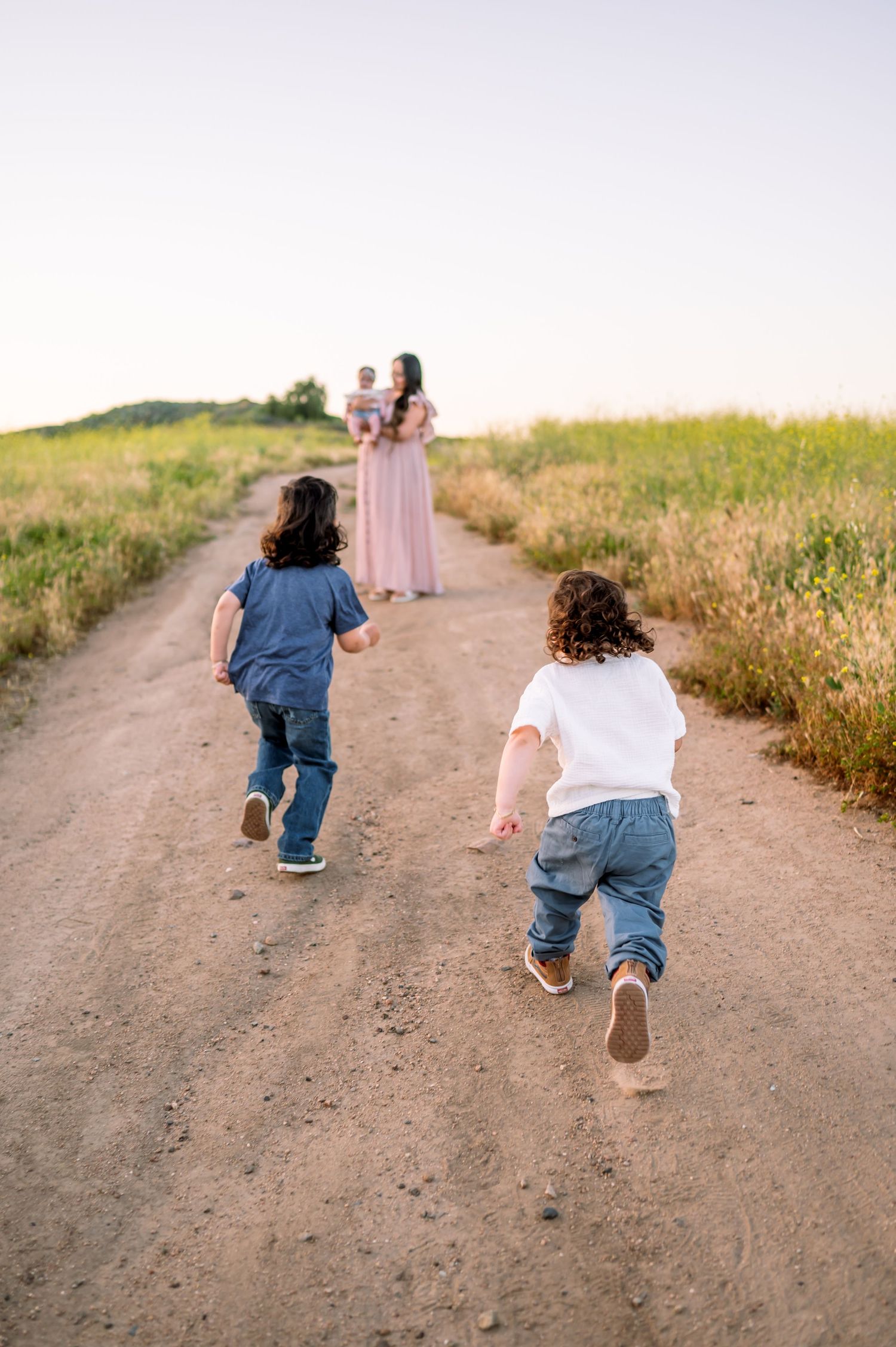 Children running towards figure in pink dress on dirt path at sunset.