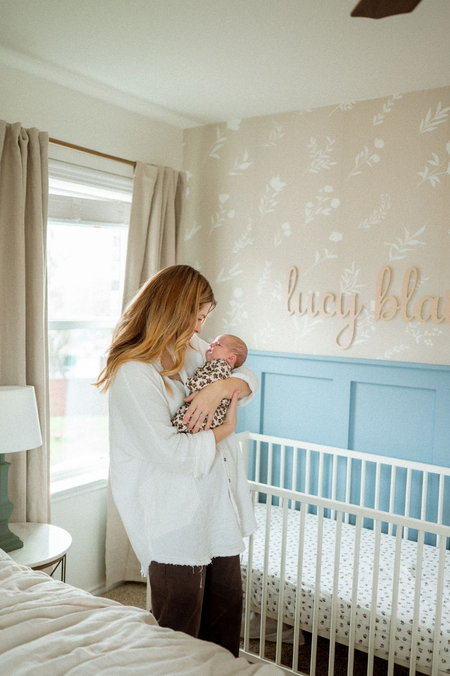 Mother holding newborn baby in nursery with floral wallpaper and blue crib.