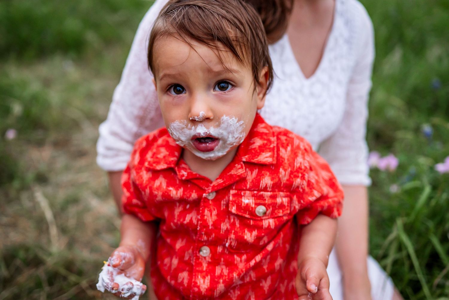 Small child in red patterned shirt with messy face sits outdoors in grass.