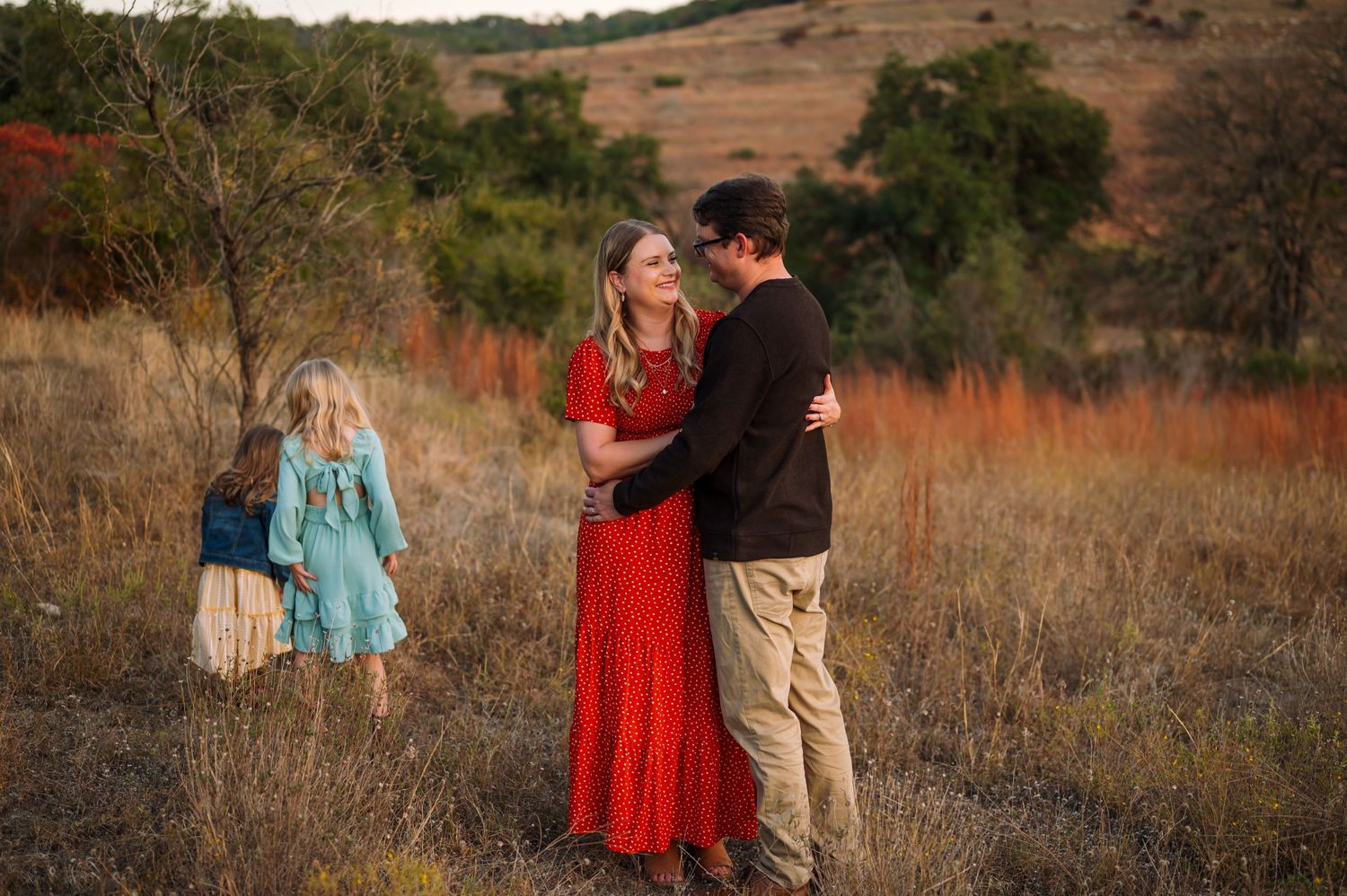 A couple shares a moment in a field while a child plays in the background.
