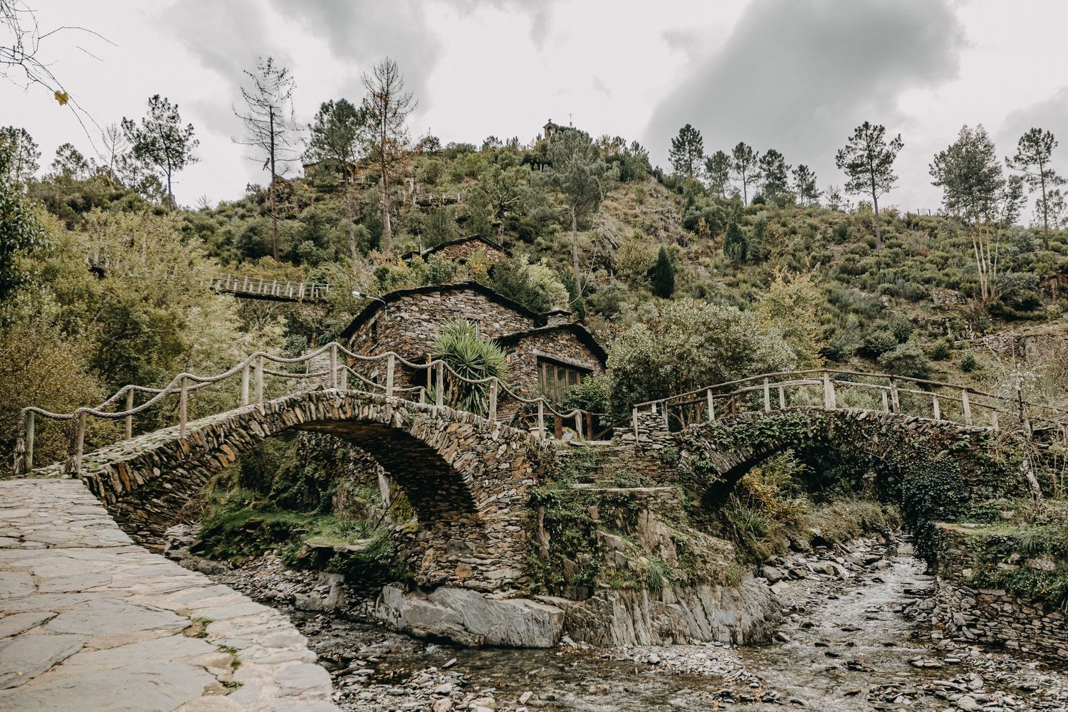 Rustic stone arch bridge crosses a rocky stream in a lush mountainous landscape.