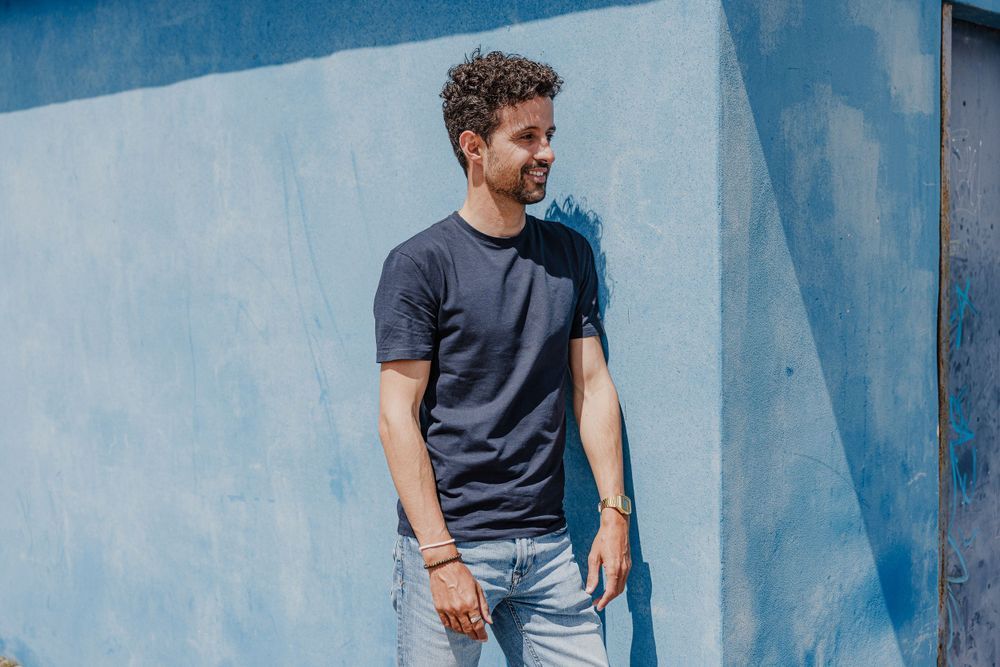 Casual portrait against blue wall wearing dark t-shirt and jeans with natural lighting.