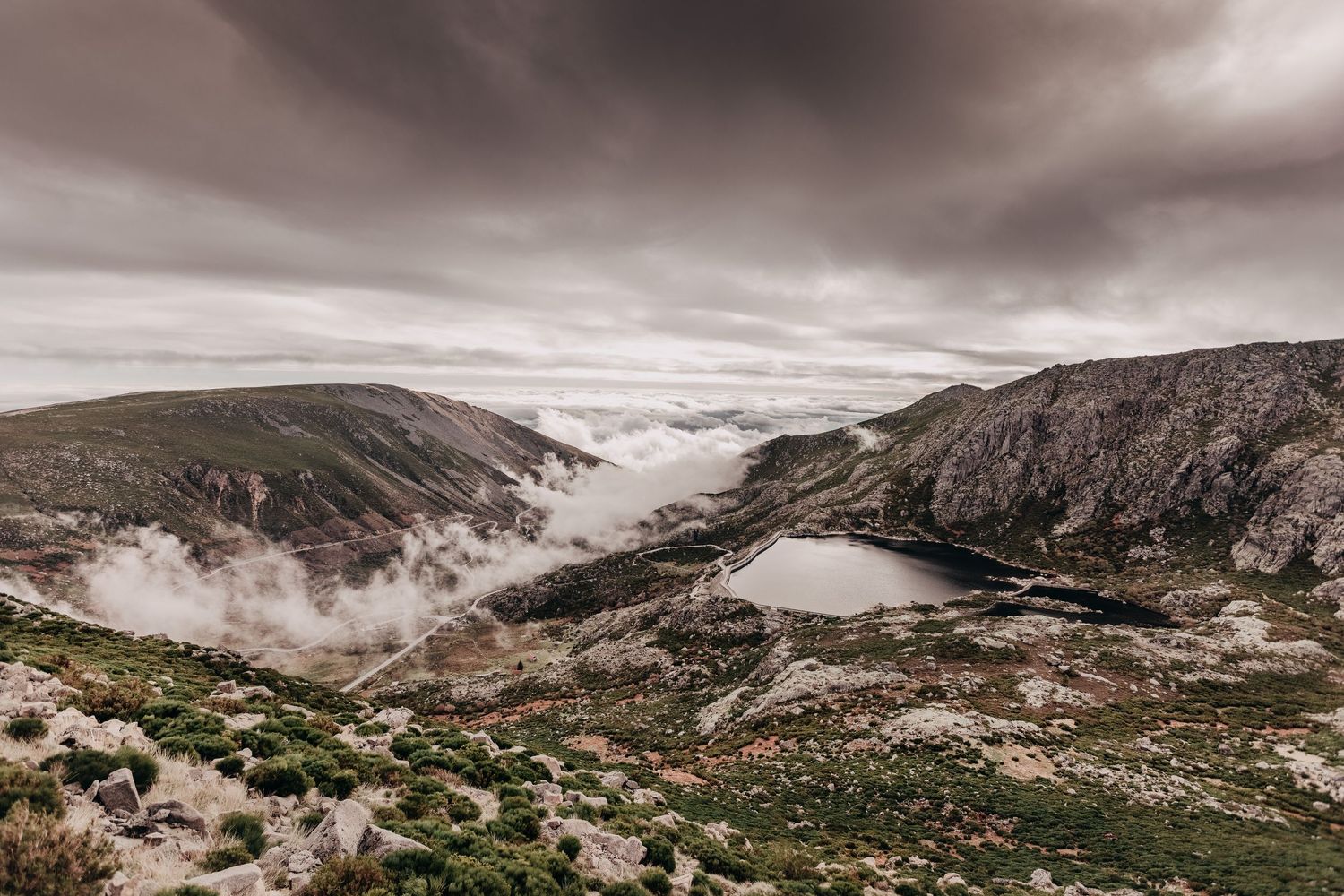 Dramatic mountain landscape with fog rolling through valley and dark lake under stormy skies.