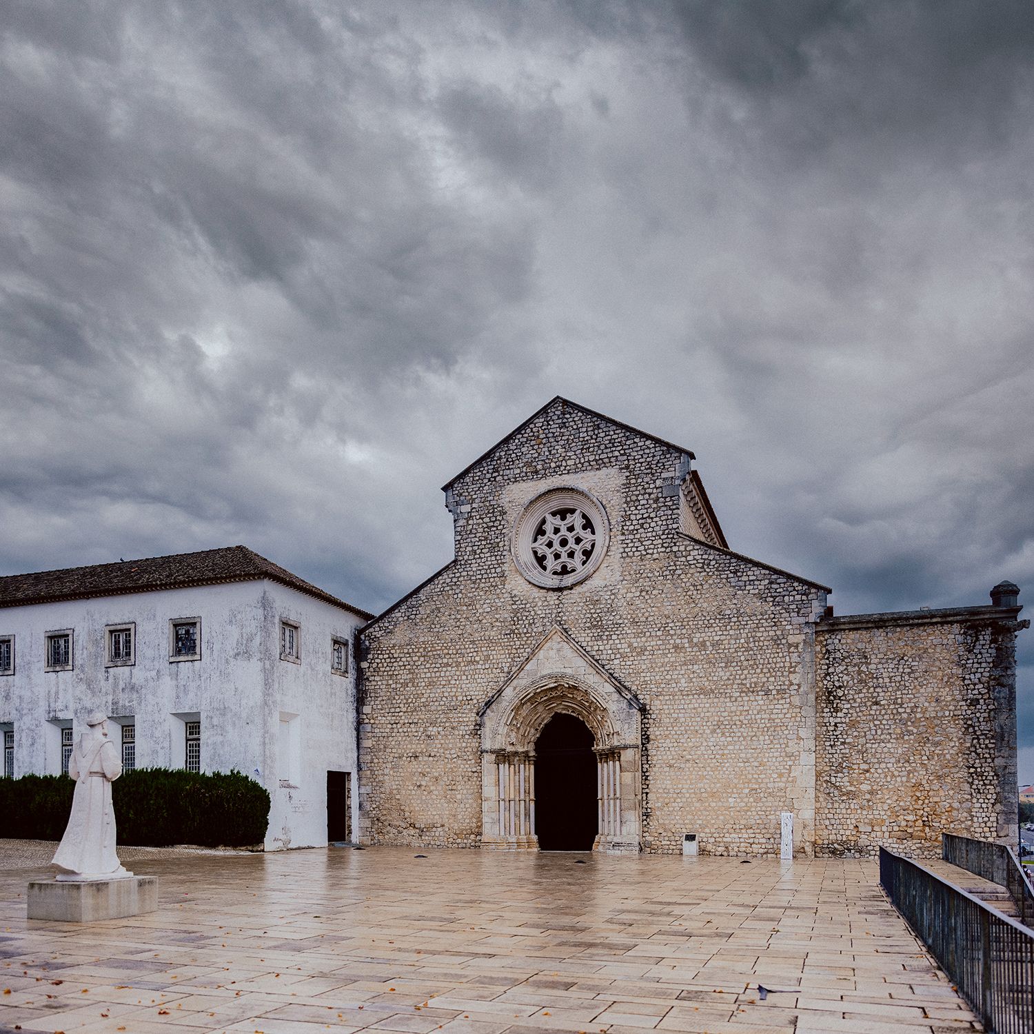 Ancient stone church with round rose window and arched entrance under dramatic stormy clouds in Portugal.