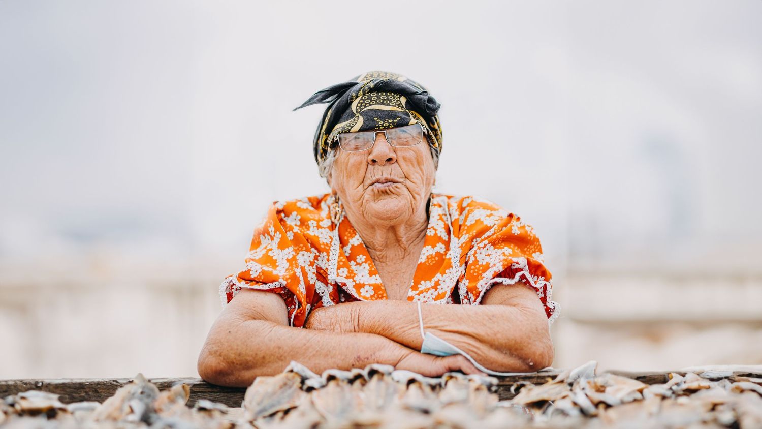 Portrait of an elder wearing an orange floral shirt and headband against a bright background.