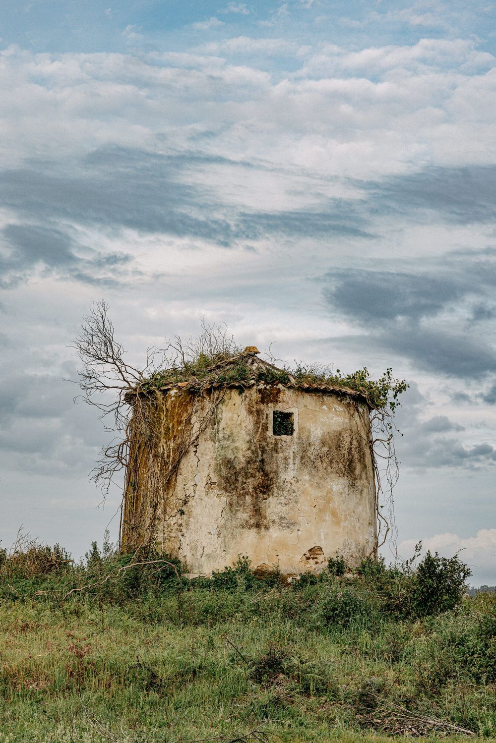 Abandoned stone tower structure on hilltop with overgrown vegetation against cloudy sky.