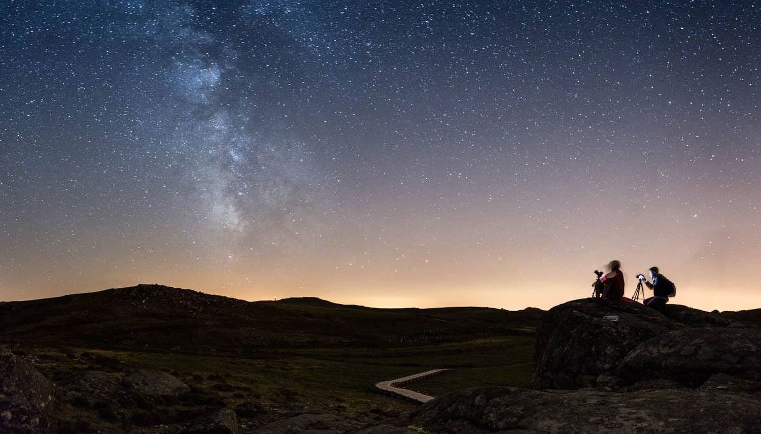 Silhouettes against starry night sky with visible Milky Way over dark rolling hills.
